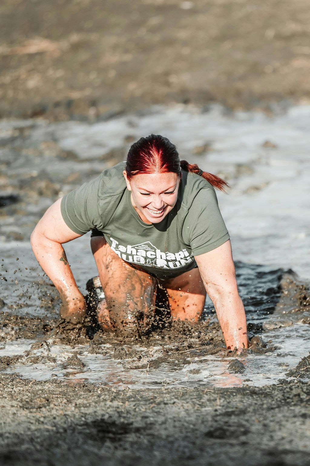 Person crawling through muddy water, smiling, wearing a green shirt.