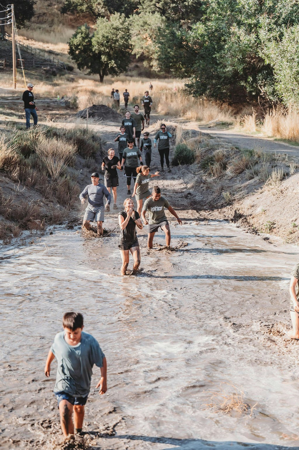People running through a muddy water obstacle in an outdoor setting with trees and grass.