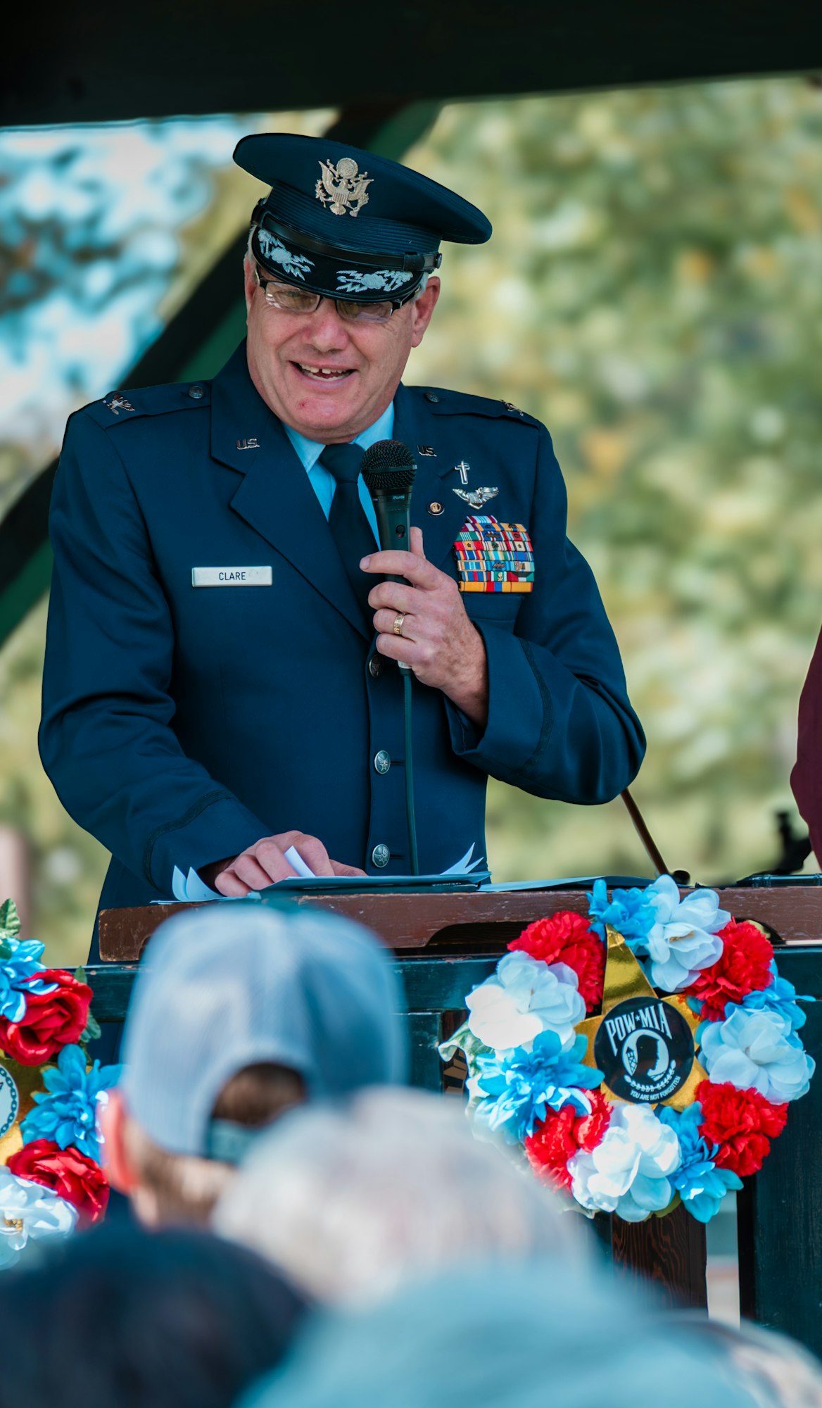 A military officer in uniform speaks at a podium with colorful wreaths, holding a microphone.
