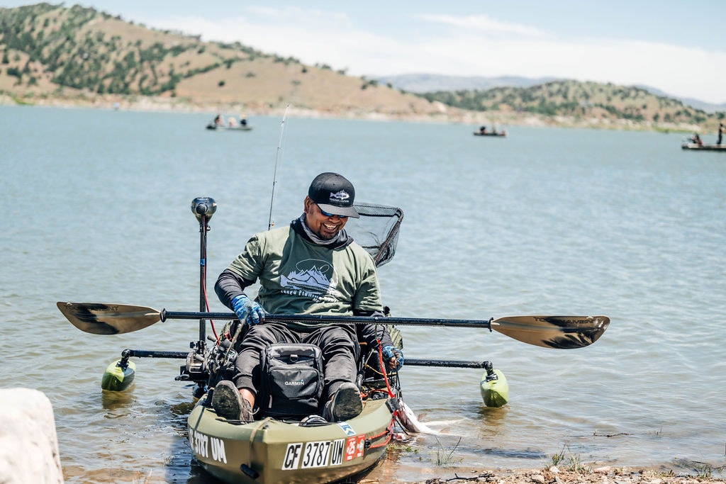 A smiling person in a kayak with fishing gear on a sunny day near the shore, with other boats in the background.