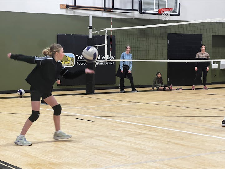 A girl playing volleyball in a gym, with other players and onlookers present.