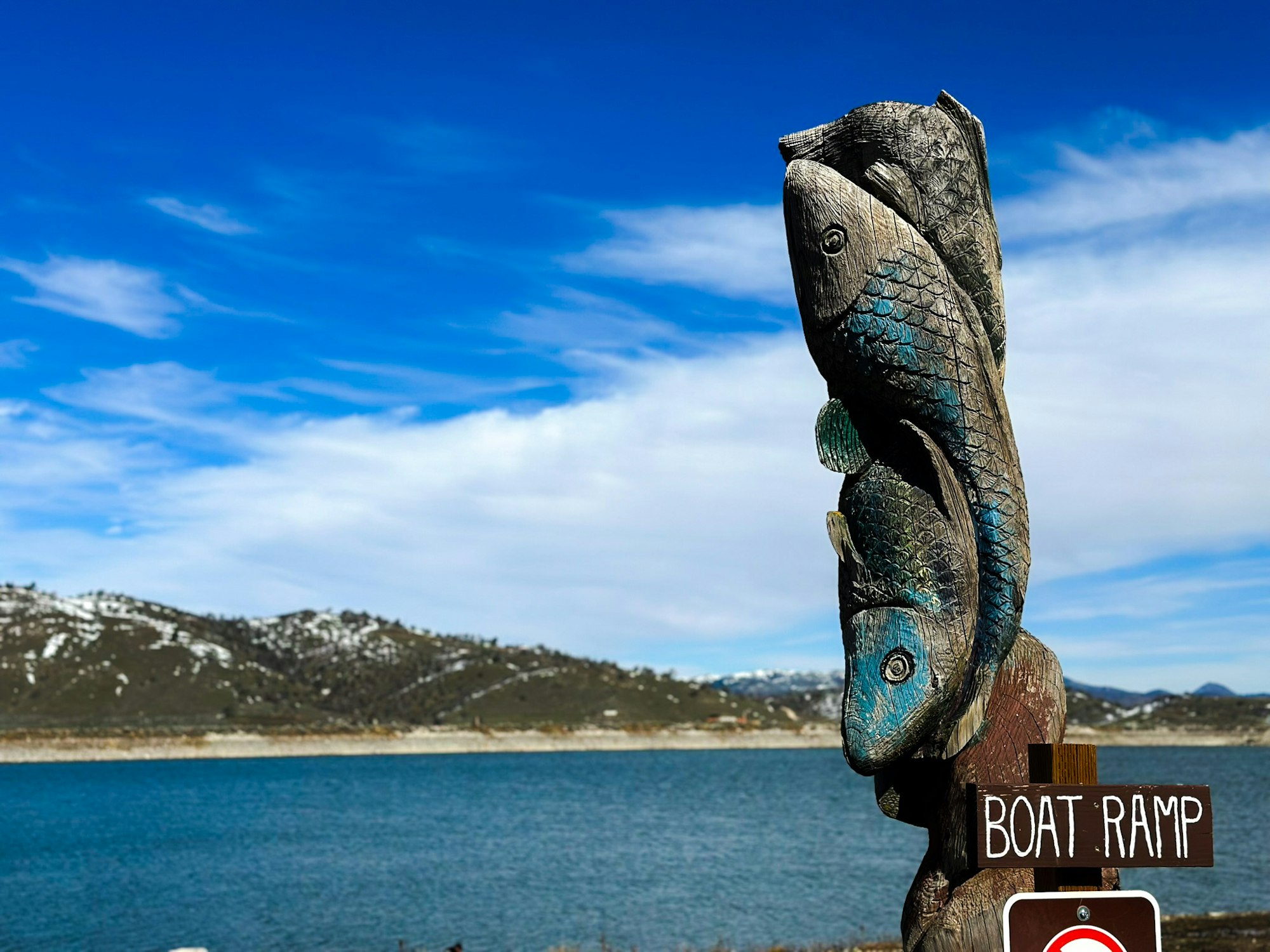 Carved wooden fish sculpture by a lake, blue sky, hill in the background, and a "Boat Ramp" sign visible.