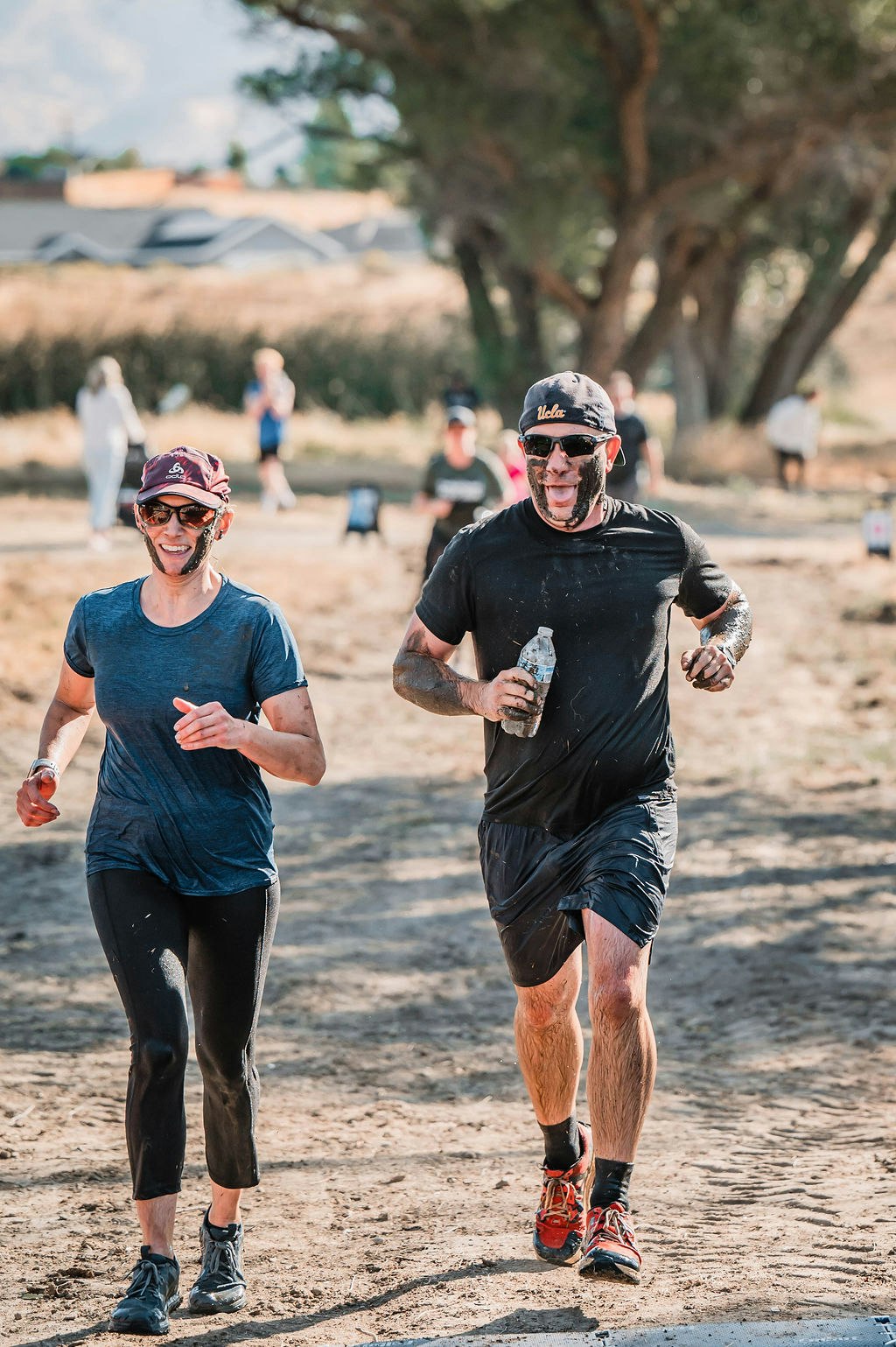 Two people, covered in mud, running outdoors on a dirt path with trees in the background.