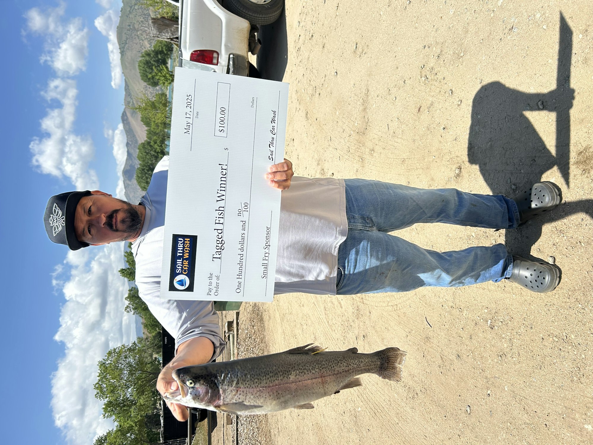 Man holds a large fish and a prize check for $100 as a "Tagged Fish Winner" outdoors, smiling.