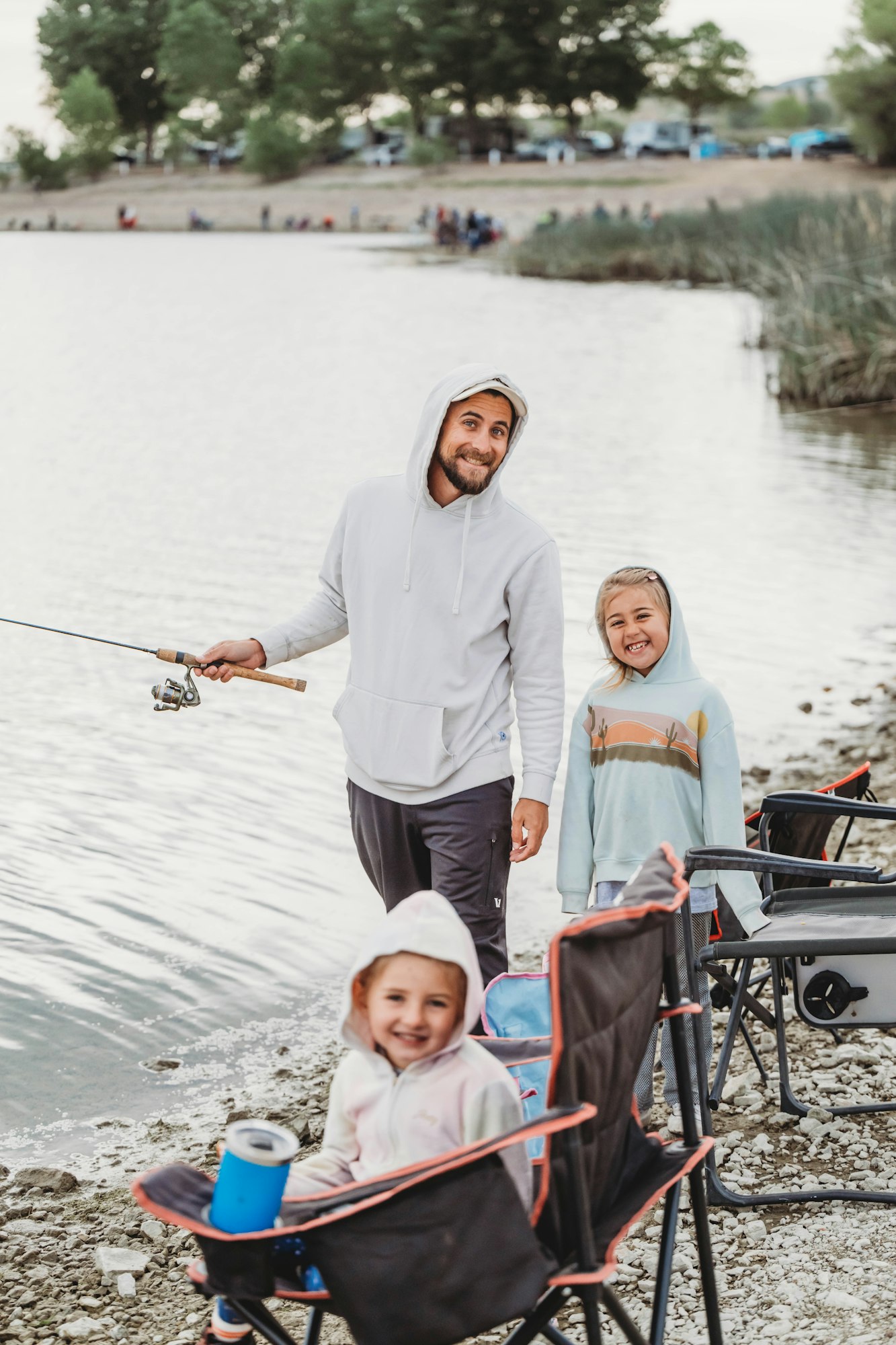 A man and two kids in hoodies by a lakeside, fishing and sitting on camping chairs.