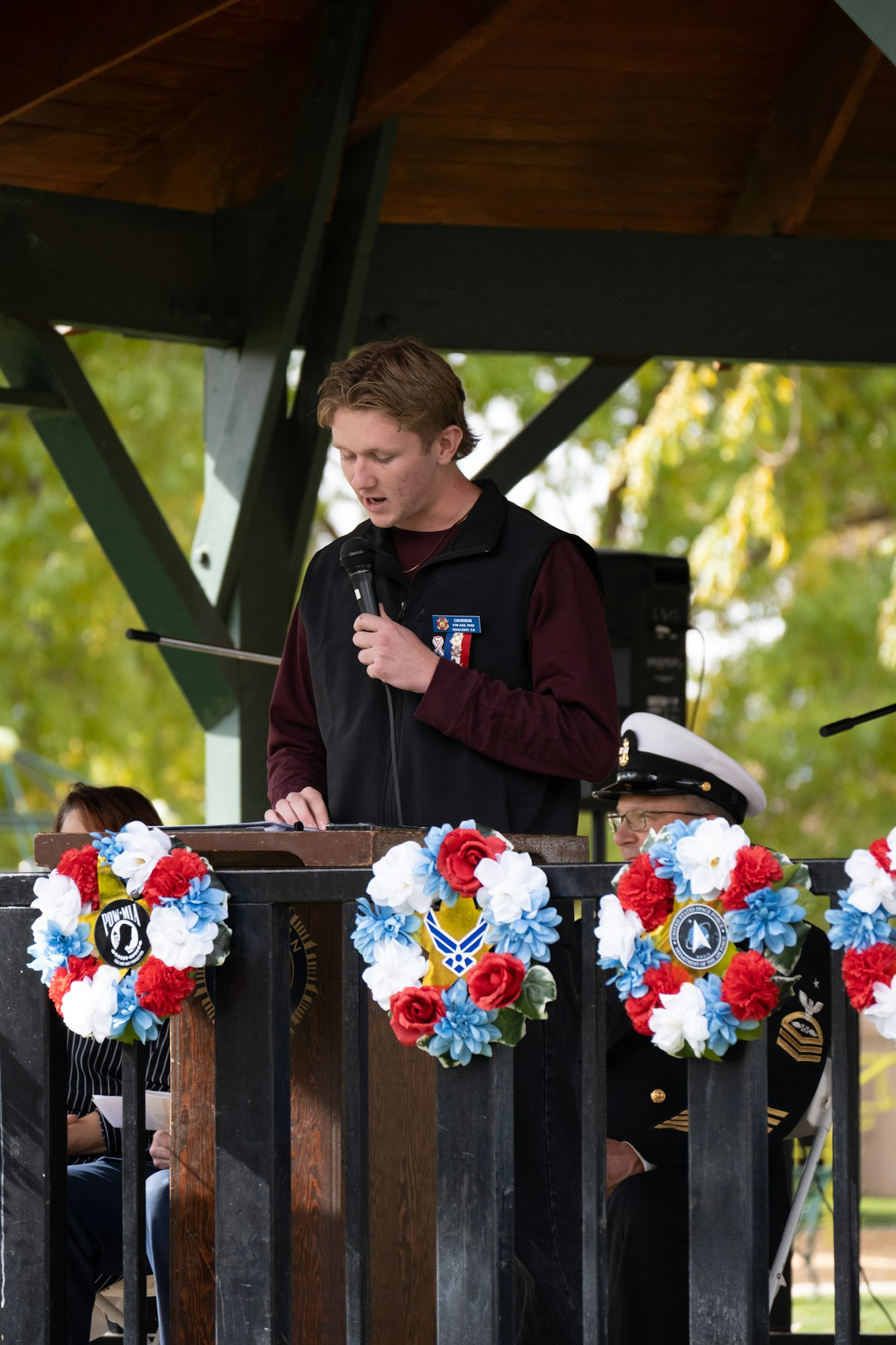 Person speaking at a podium adorned with patriotic wreaths, holding a microphone, in an outdoor setting.