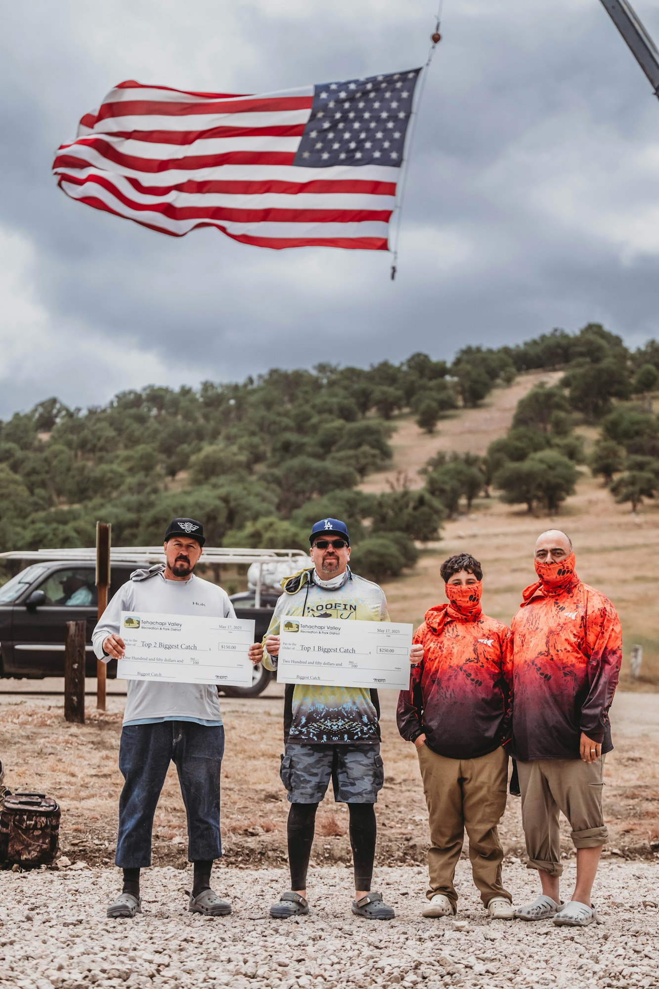 Four people posing outdoors with large checks; an American flag waves in the background.