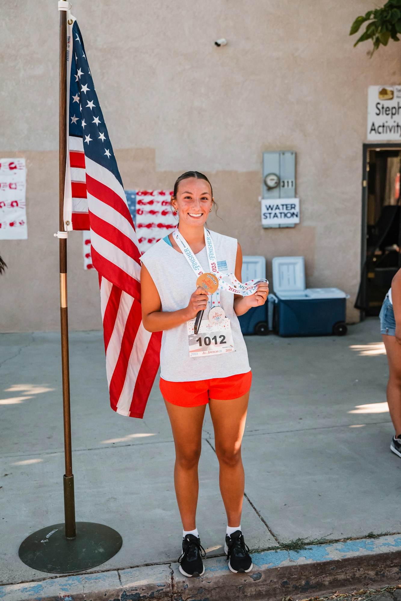 Young woman wearing a race number and medal, standing by an American flag, smiling. Signs in the background.