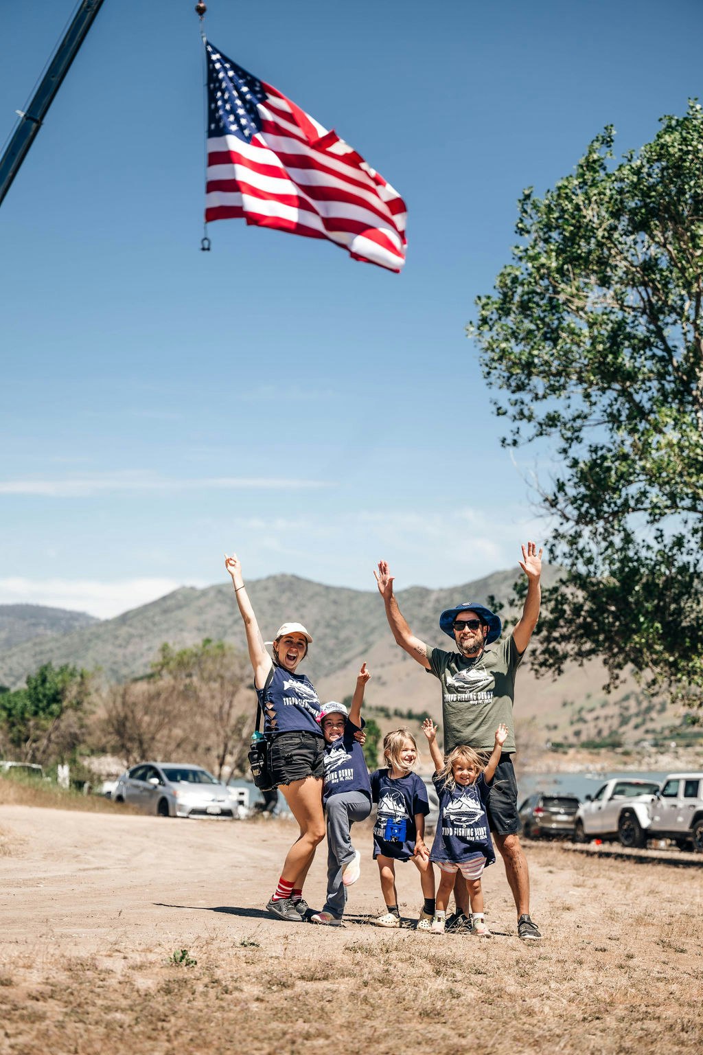 A happy family with two adults and three kids raising their hands in excitement outdoors, with an American flag in the background.