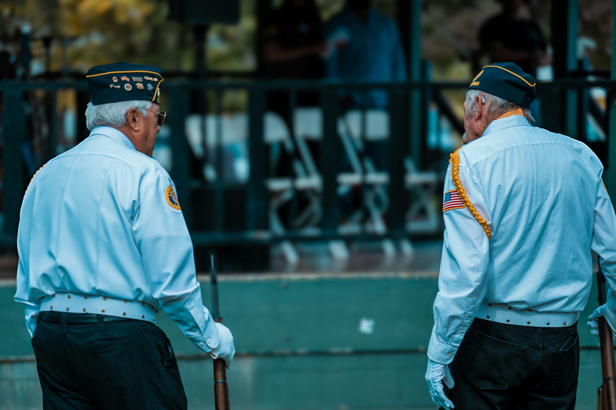 Two men in uniform, wearing hats with badges, stand outdoors holding rifles, viewed from the back.