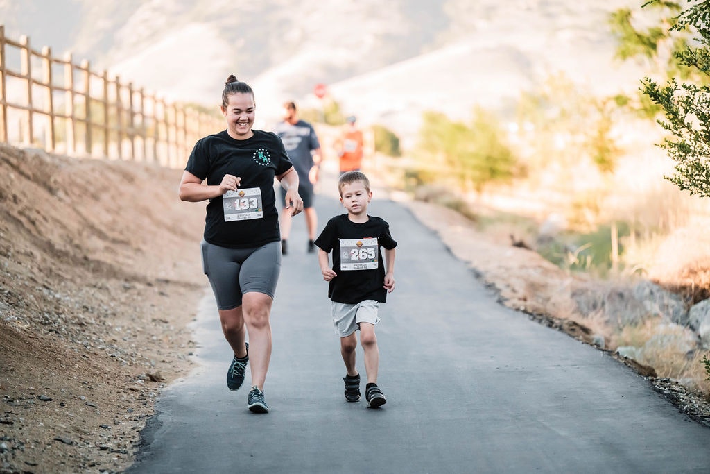 A woman and a child are jogging on a path wearing race numbers, with a scenic backdrop.