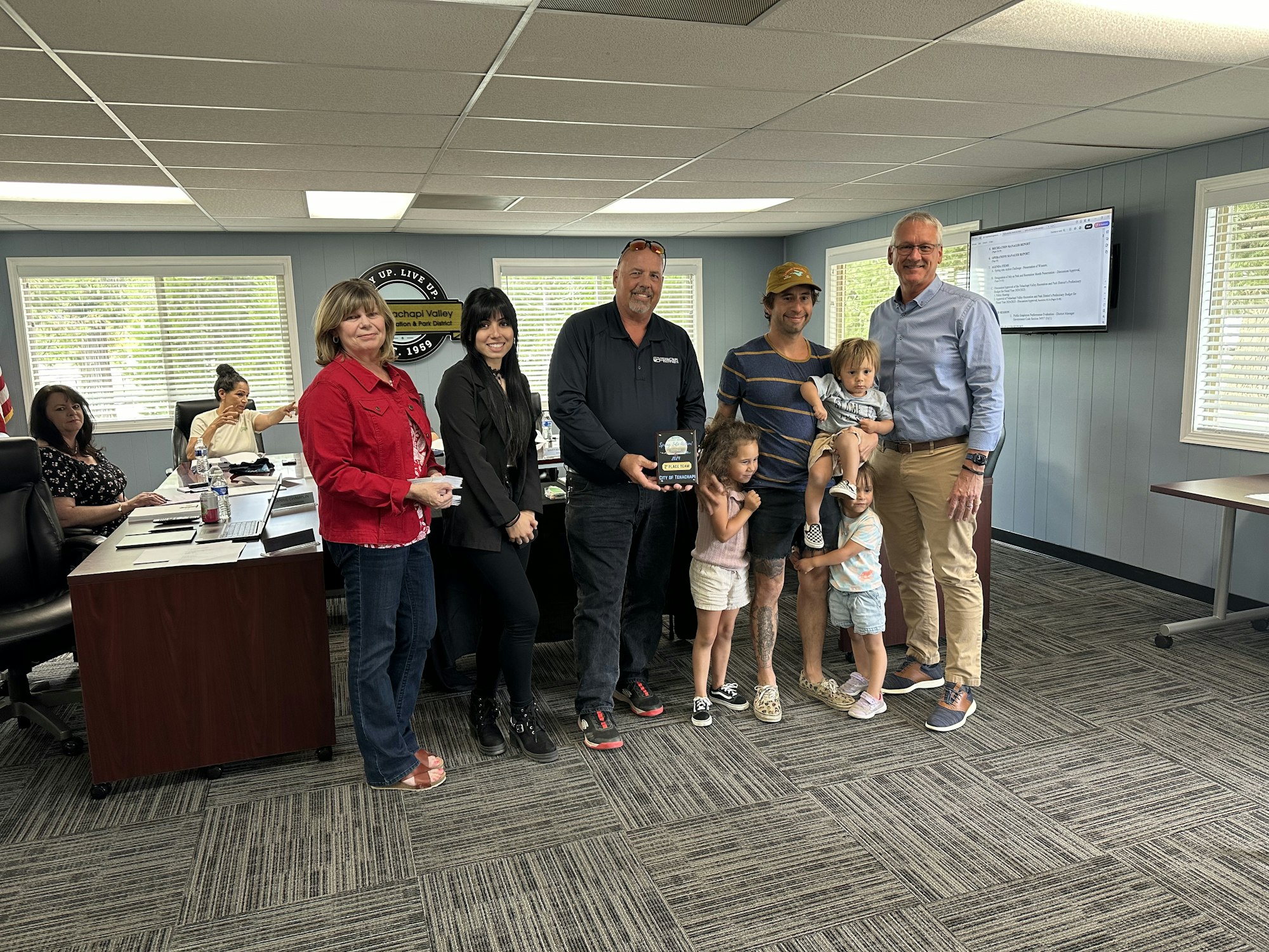 A group of adults and children in an office, one holding an award.