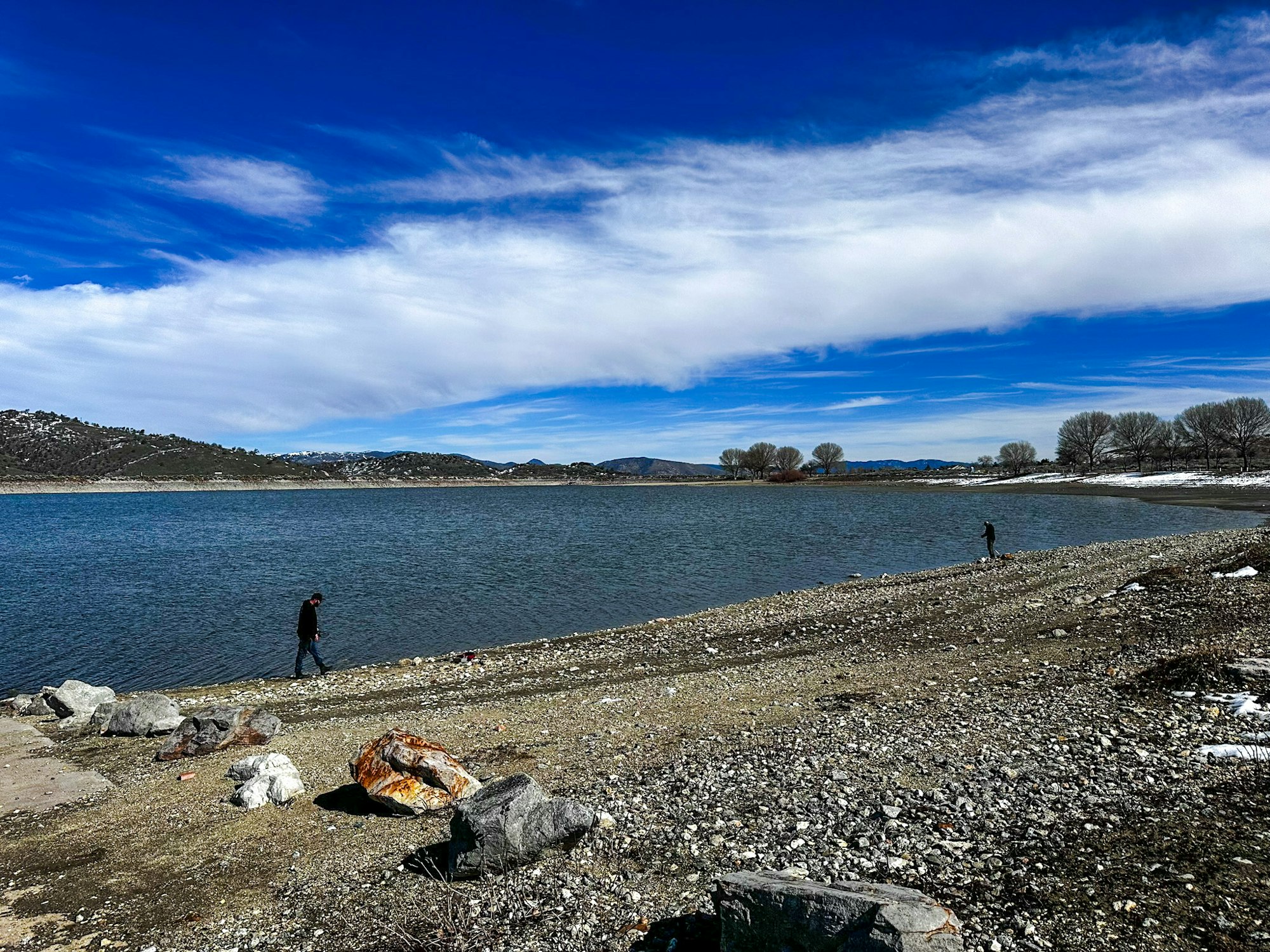 A lakeside landscape with two people, blue sky, clouds, and patches of snow on the ground.