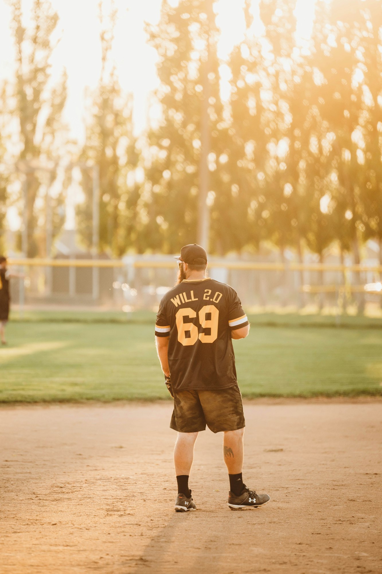 A person wearing a jersey stands on a sunlit baseball field, viewed from behind.