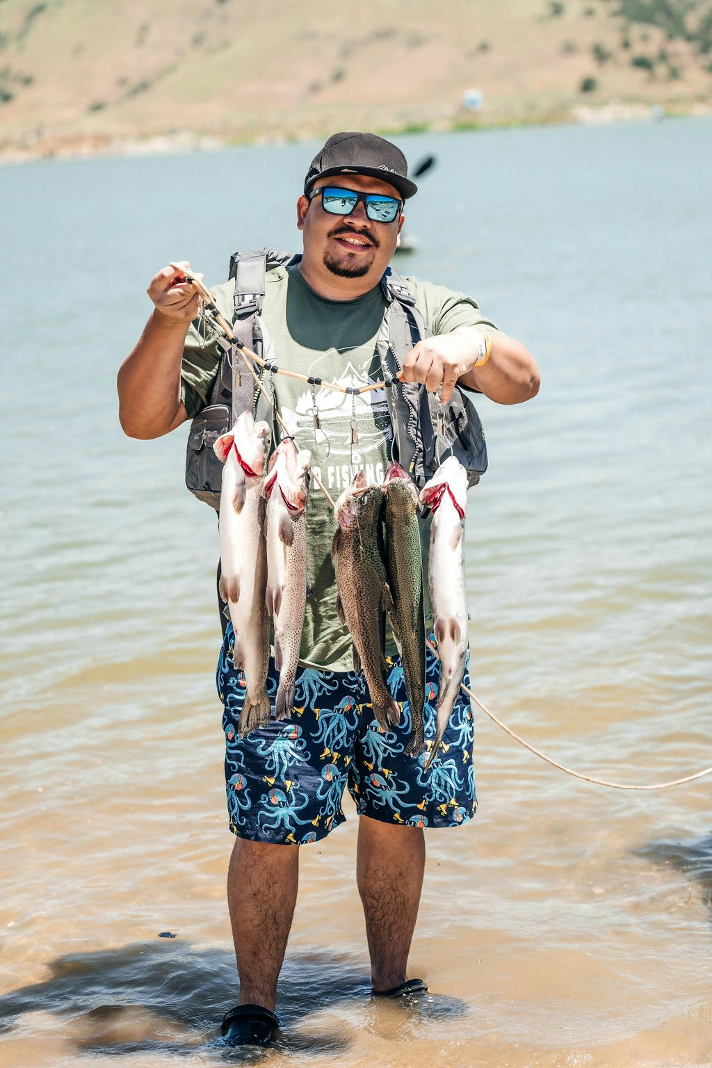 A man standing in water holding a string of fish, wearing a hat, sunglasses, and a life vest.