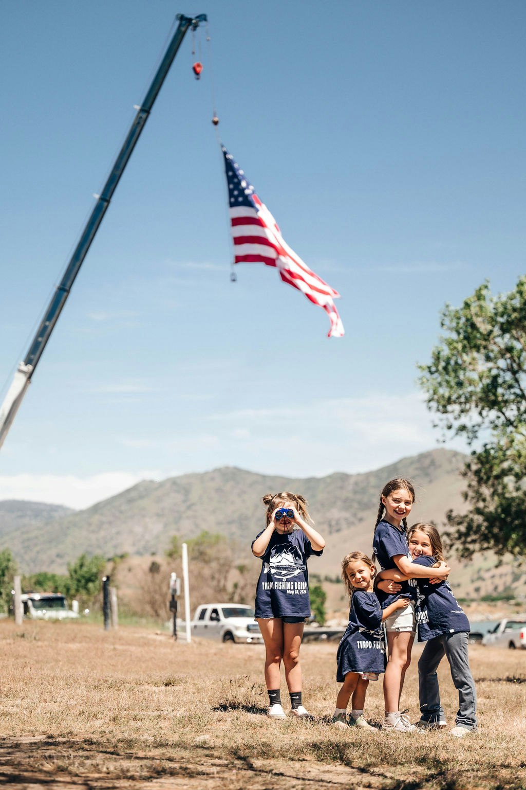 Four children outside, one looking through binoculars, an American flag hoisted by a crane in the background.