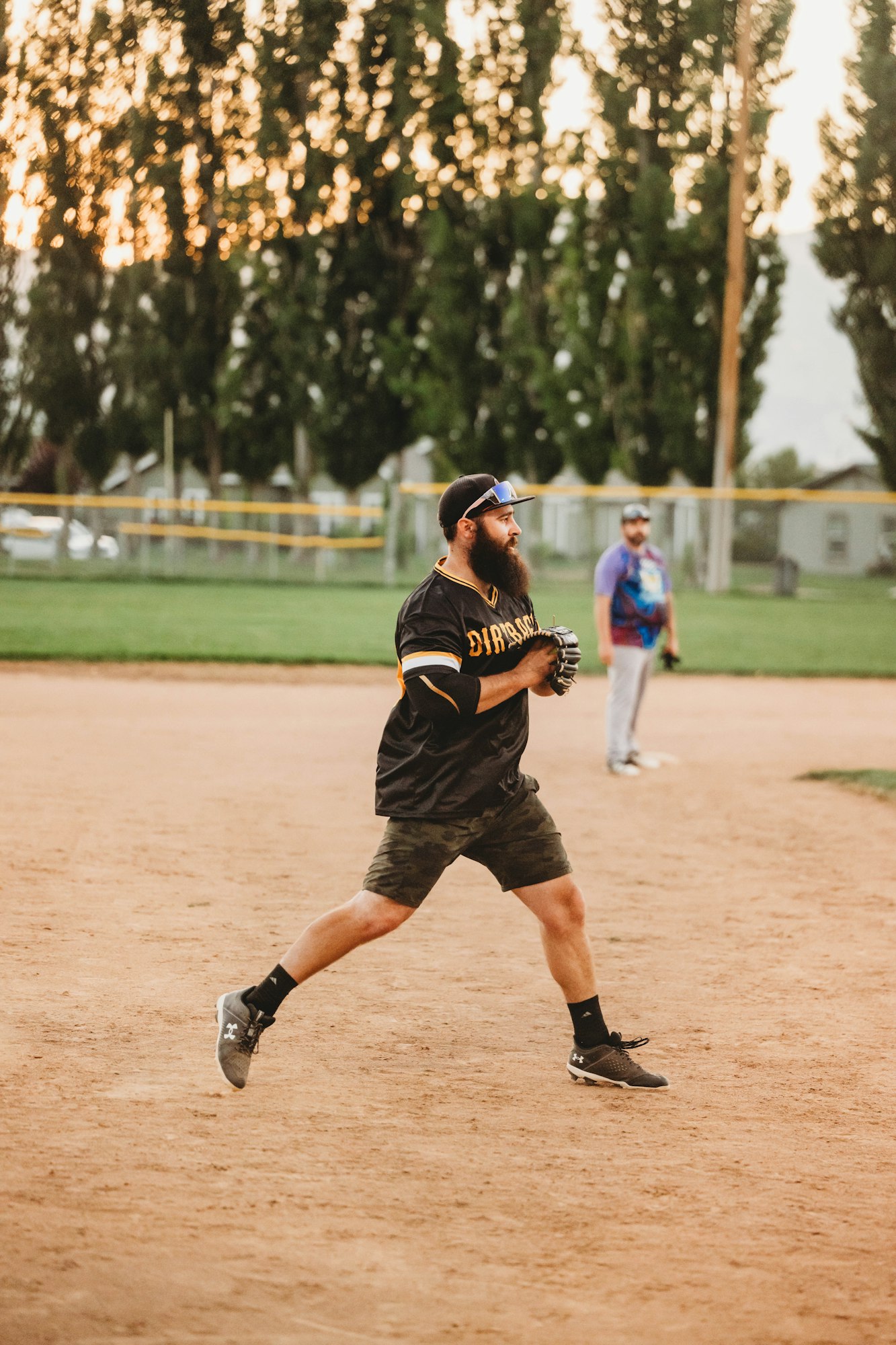 A bearded man is pitching a baseball in a field at dusk with another player in the background.