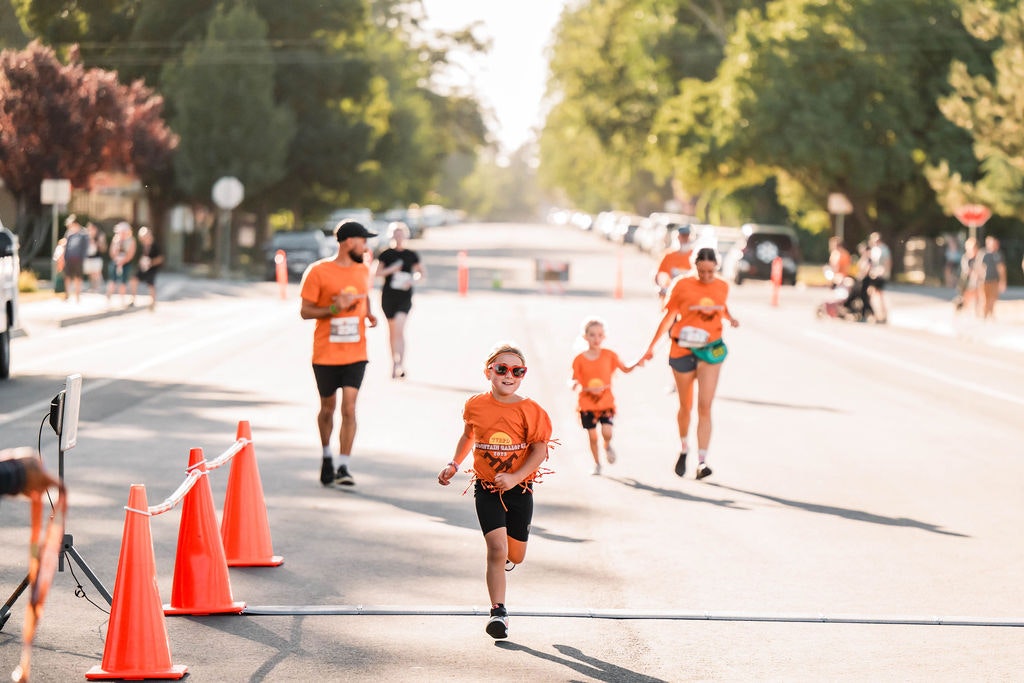 People in orange shirts, including a child, running towards the finish line of a race on a sunny street.