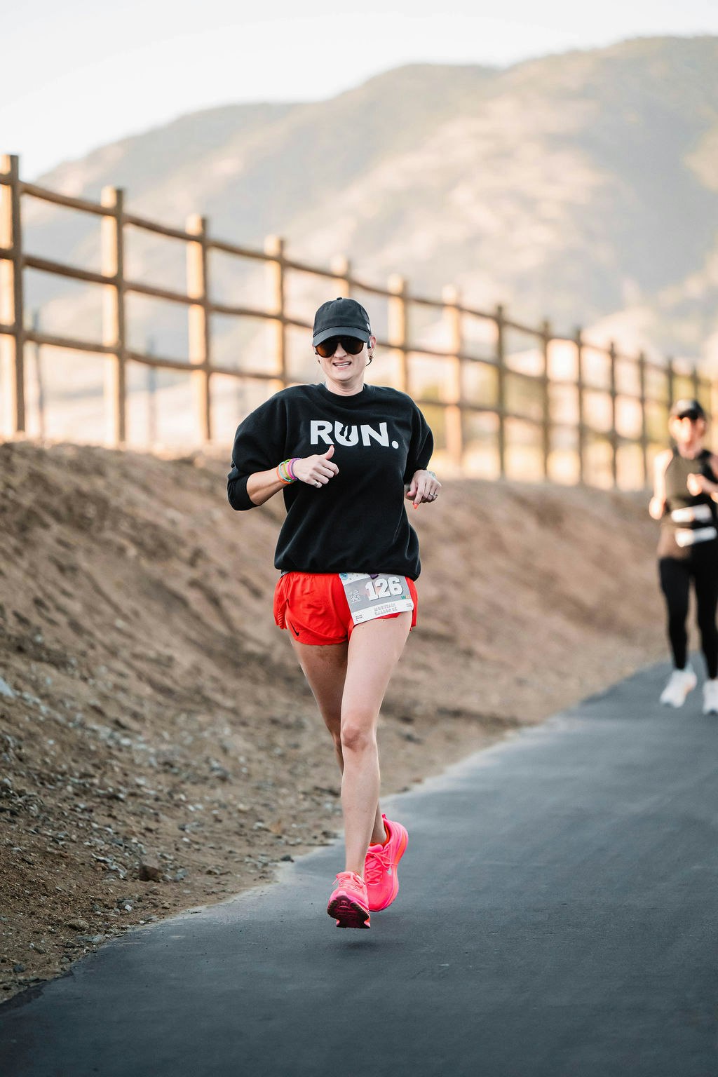 Runner in red shorts and pink shoes on a trail, wearing a "RUN." shirt, black cap, and sunglasses, with a number bib.
