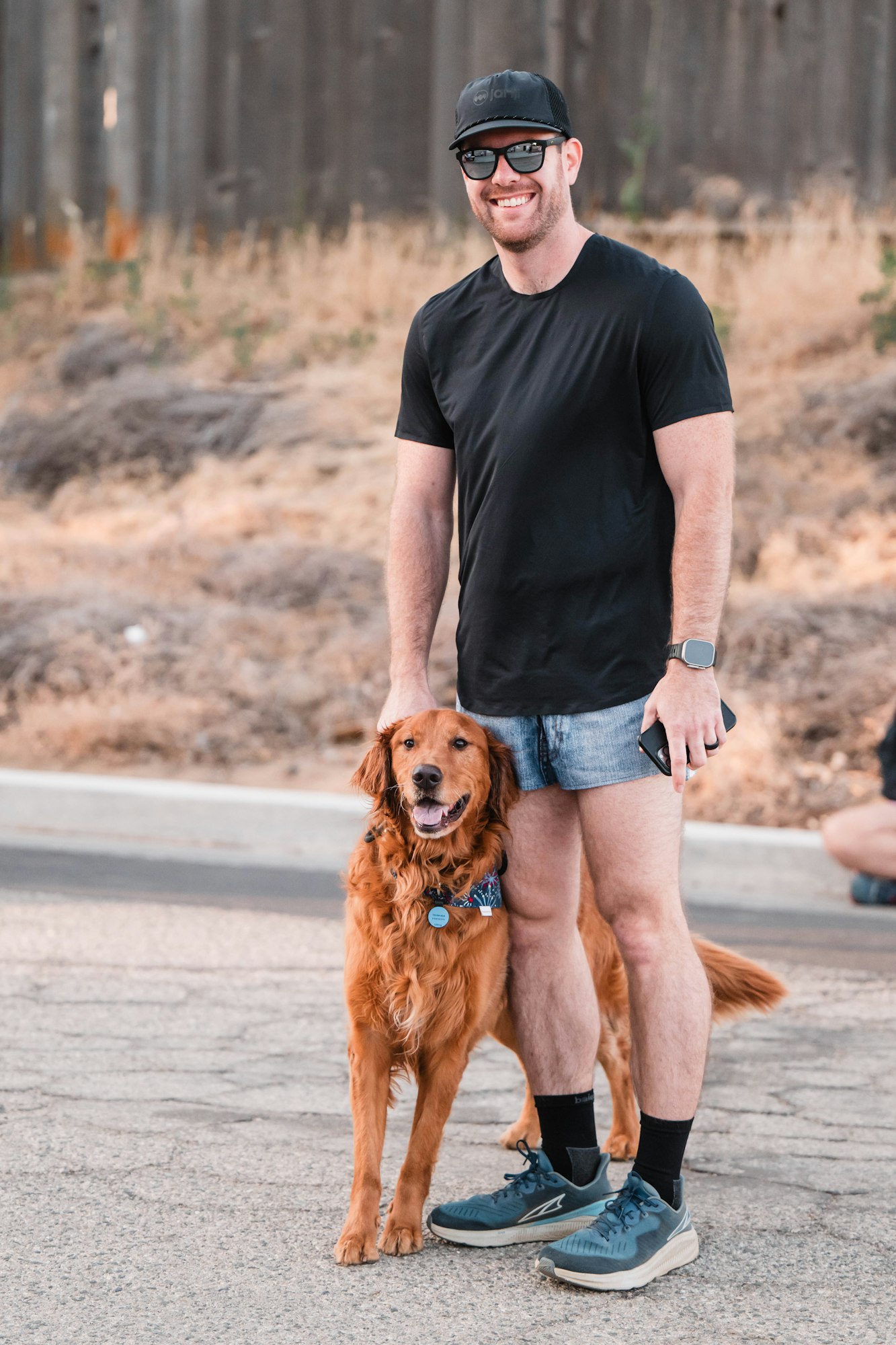 A smiling person in sunglasses and shorts standing with a golden retriever on a street.