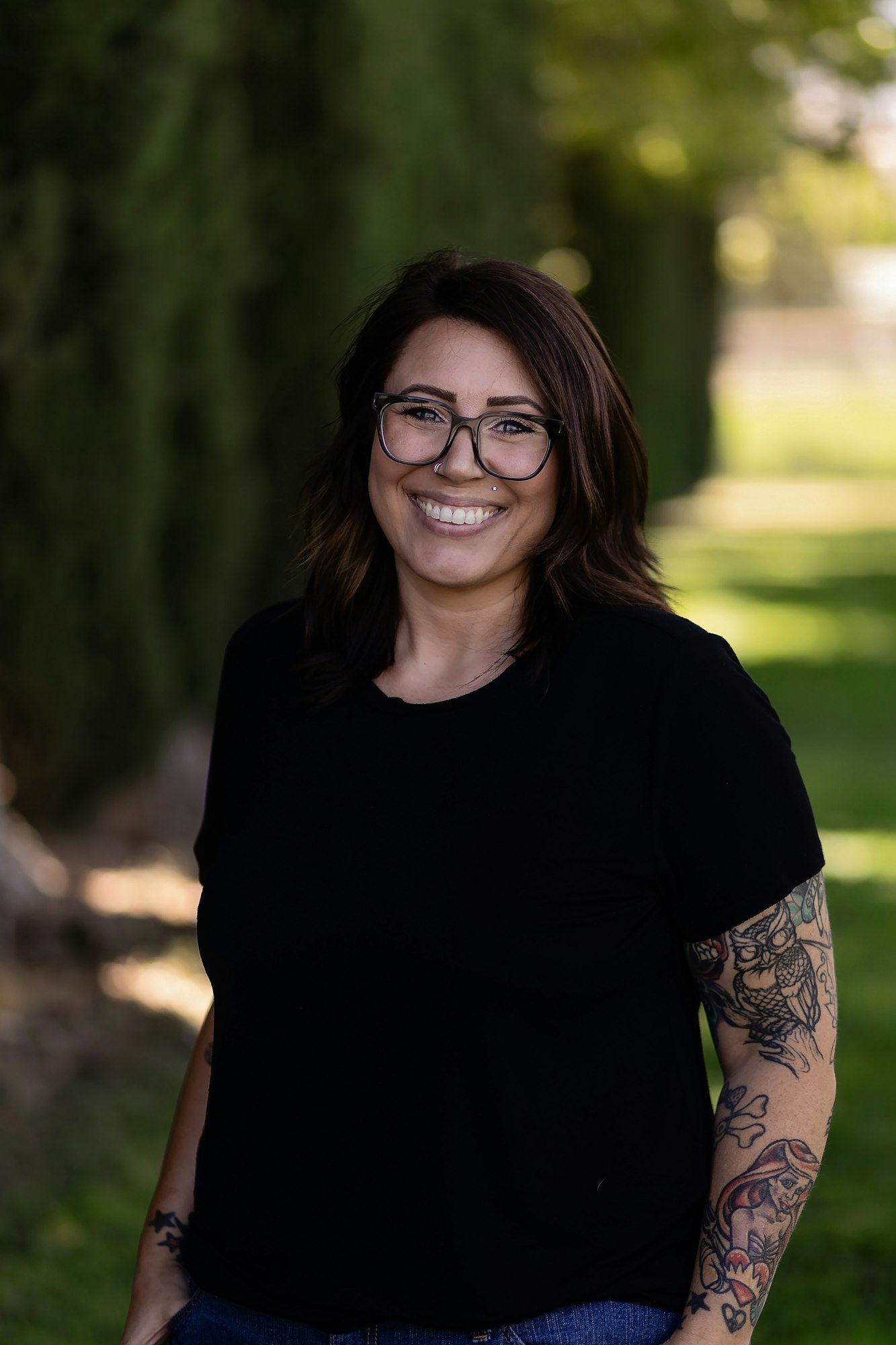 A person with glasses and tattoos stands outdoors, smiling, wearing a black shirt.