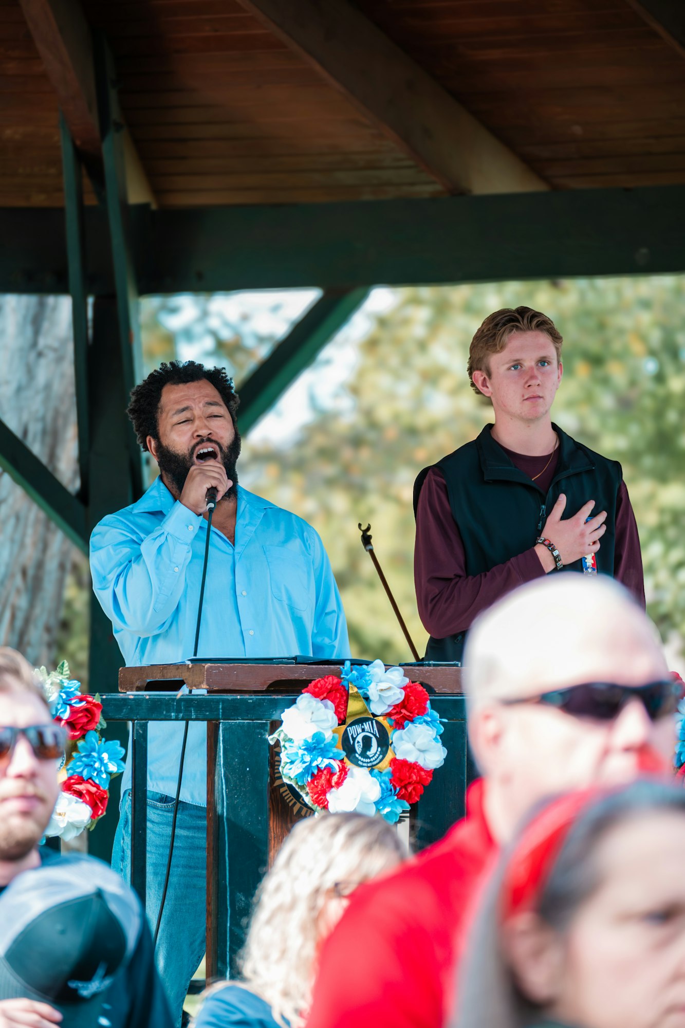 Two people on stage at a patriotic event, one singing into a microphone, the other with hand over heart, surrounded by a crowd.