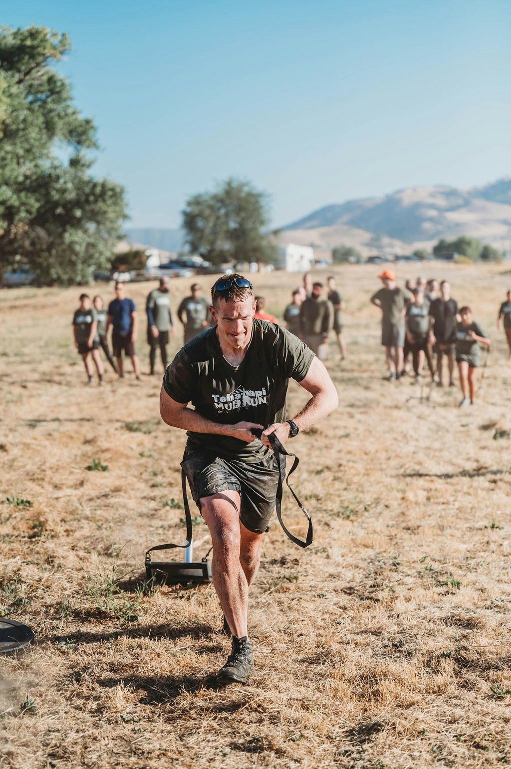 A man participating in an outdoor mud run event, pulling a weighted sled, with spectators in the background.