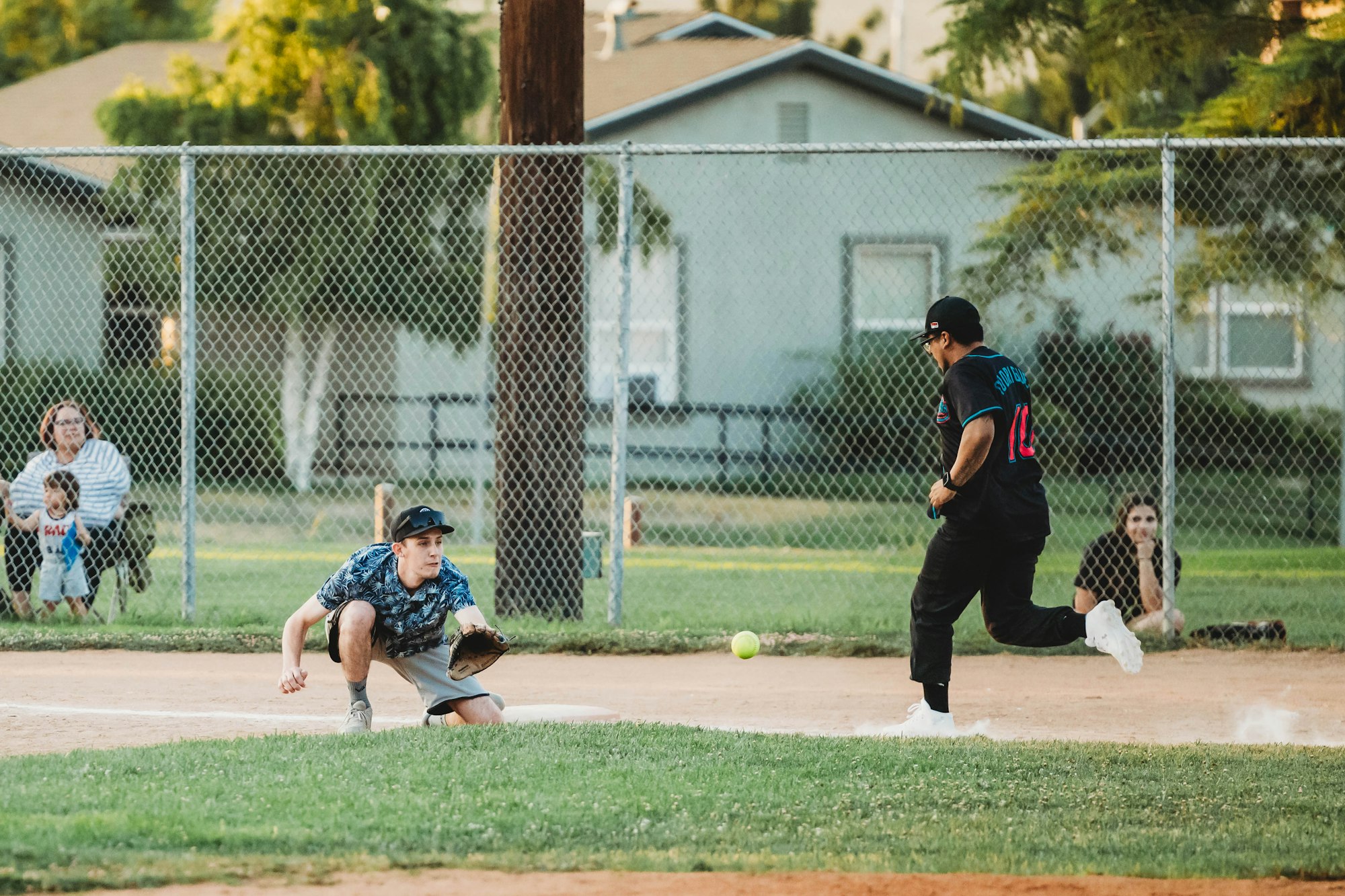 A softball game in progress with a pitcher, catcher, and spectators behind a chain-link fence.