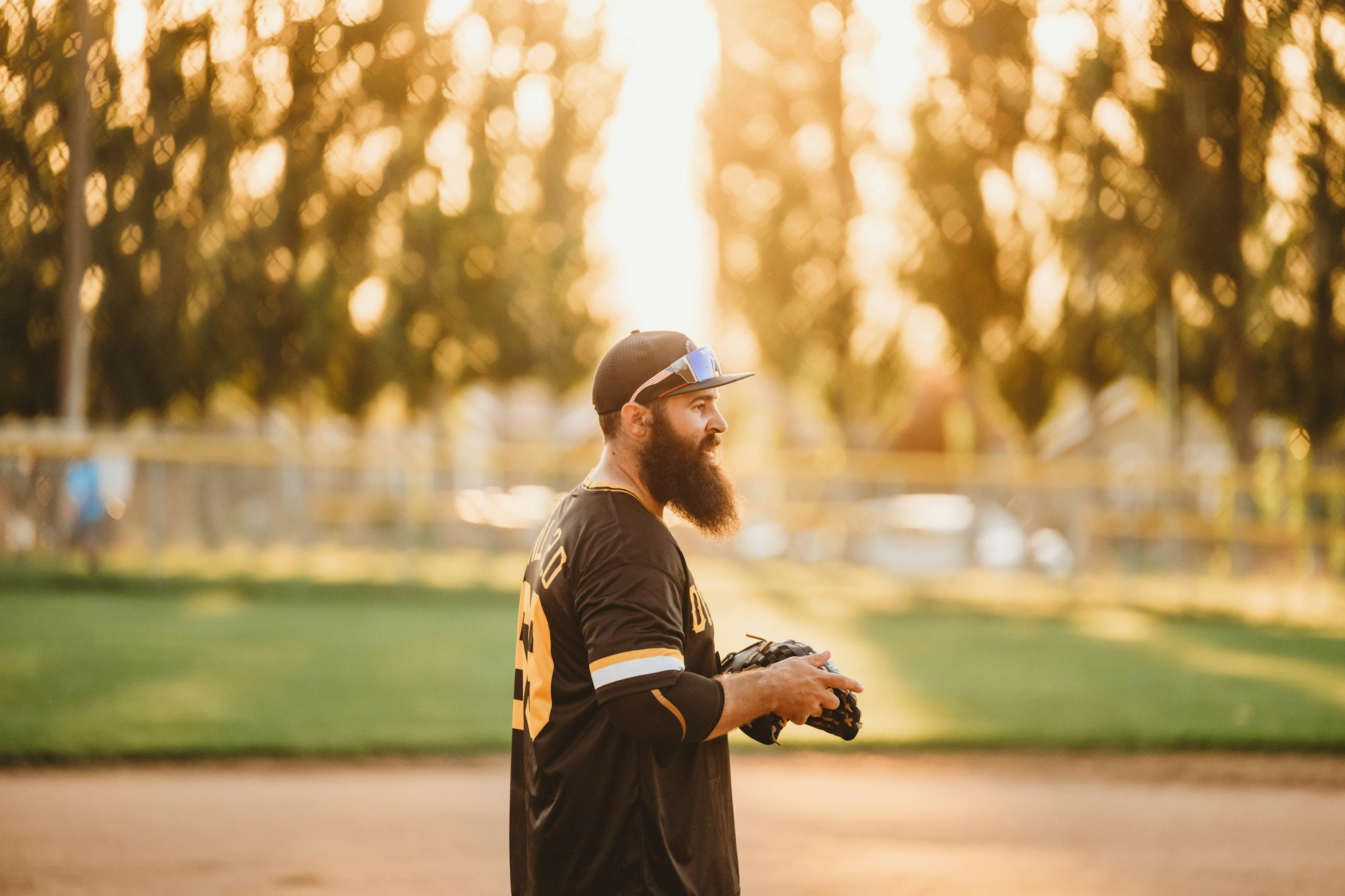 A bearded baseball player in a black and gold uniform with a glove, standing on a field at sunset.