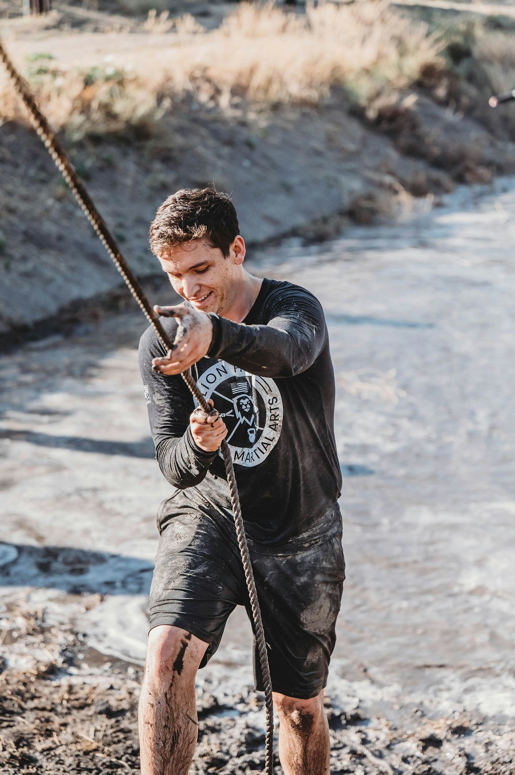 A person in muddy clothes is pulling a rope near a riverbank.