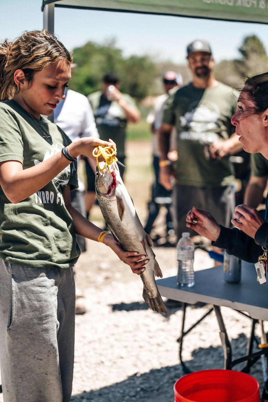 A girl shows a fish with a tag at an outdoor event to a smiling onlooker.