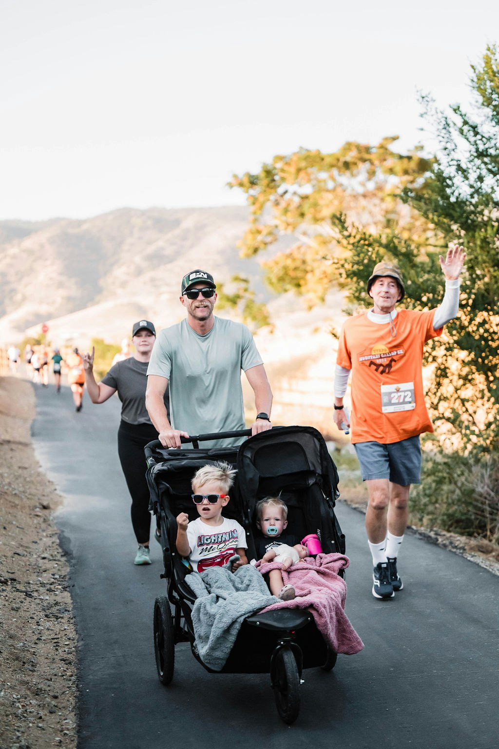 A man pushes a double stroller with two children during a race; other participants walk and wave.
