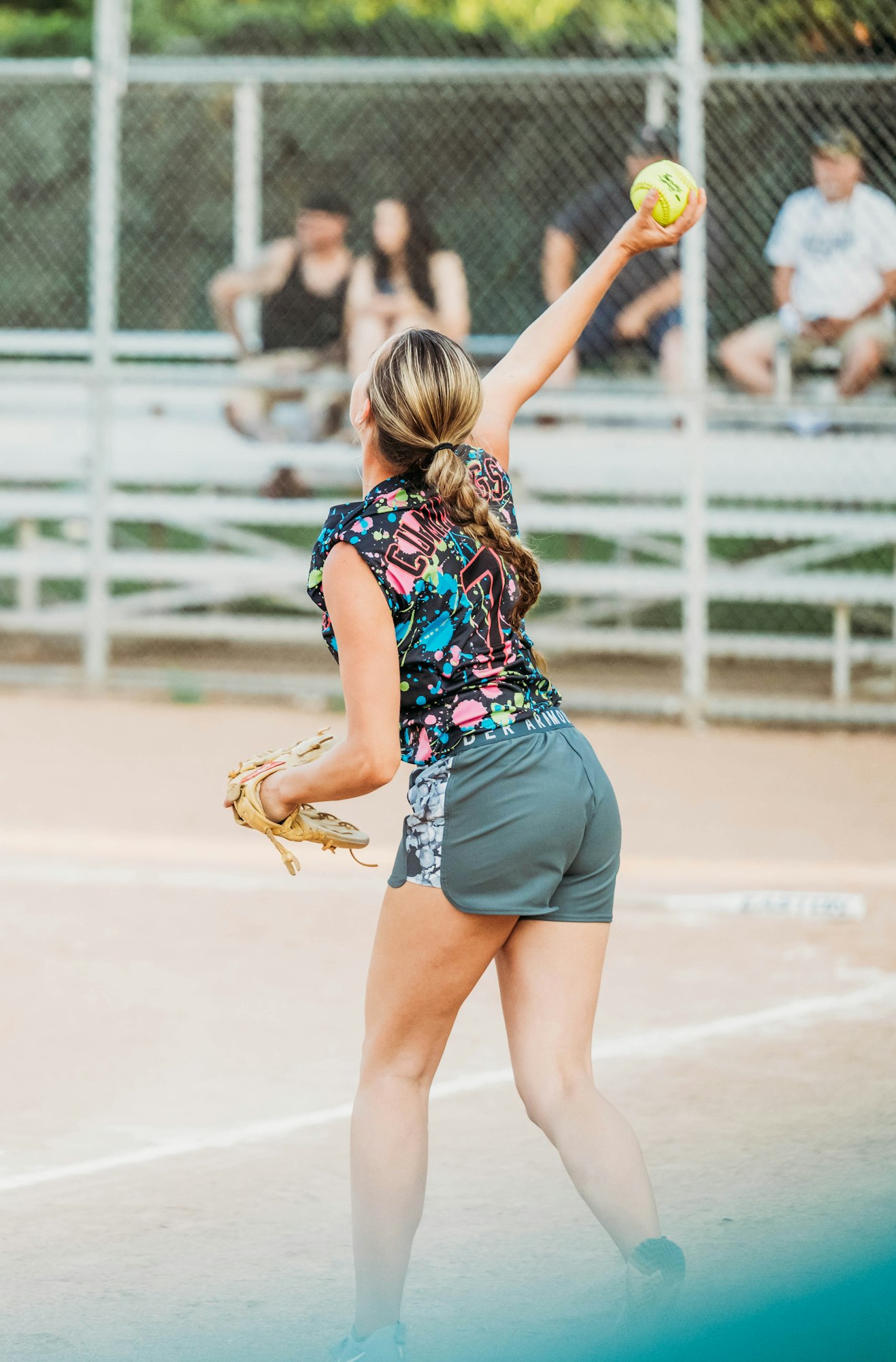 A softball player is pitching on a dirt field with spectators in the background.