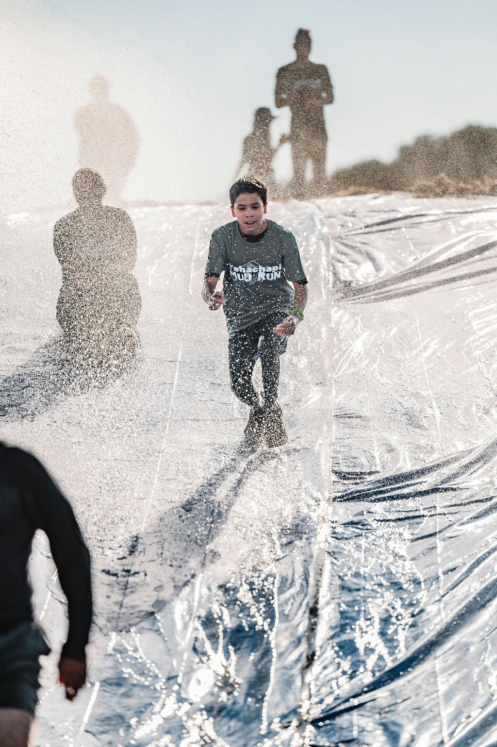 A child runs down a wet slide, splashing water, with blurred shadows of other people in the background.