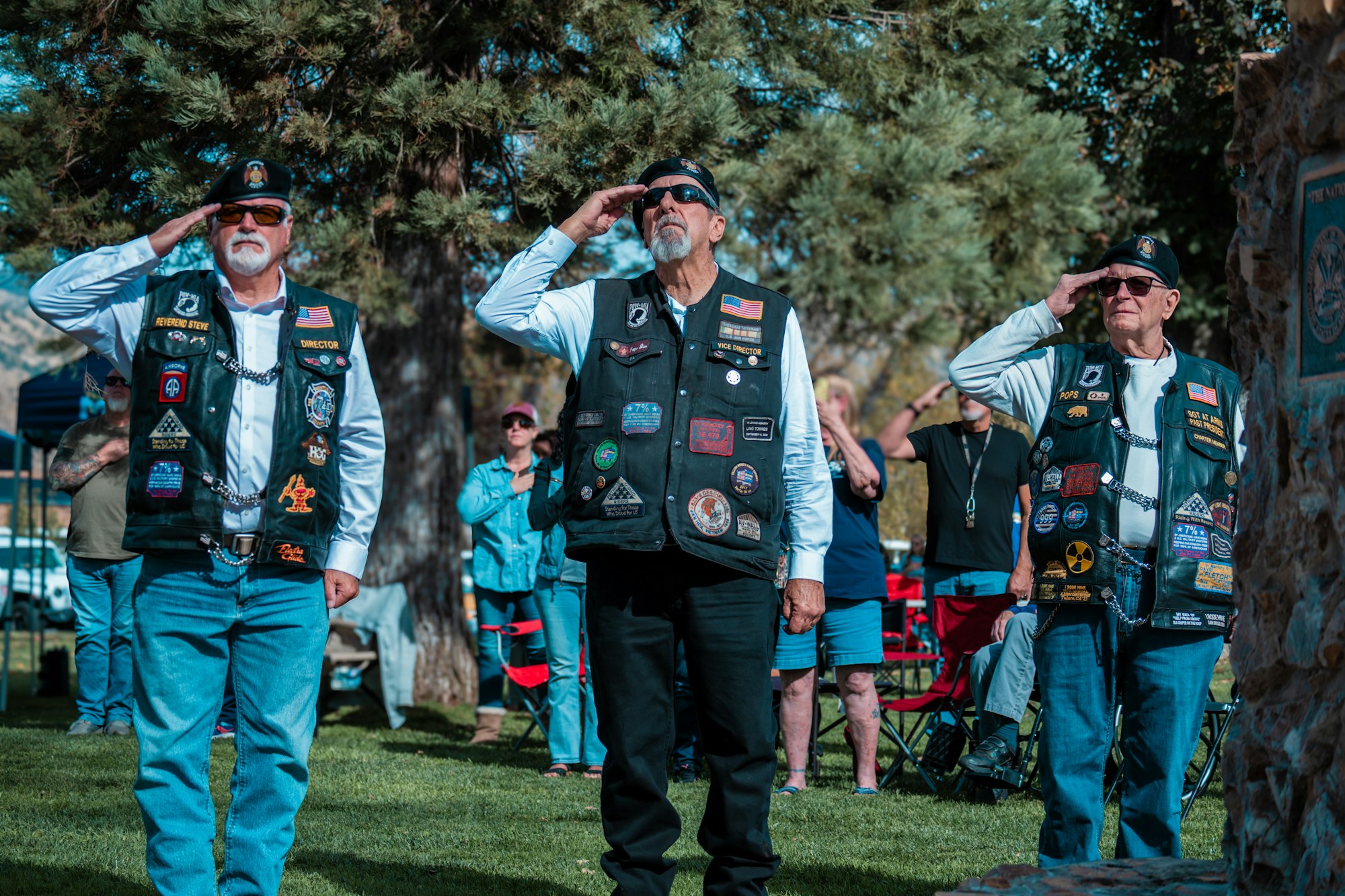 Three people in decorated vests are saluting outdoors, with a few others in the background.