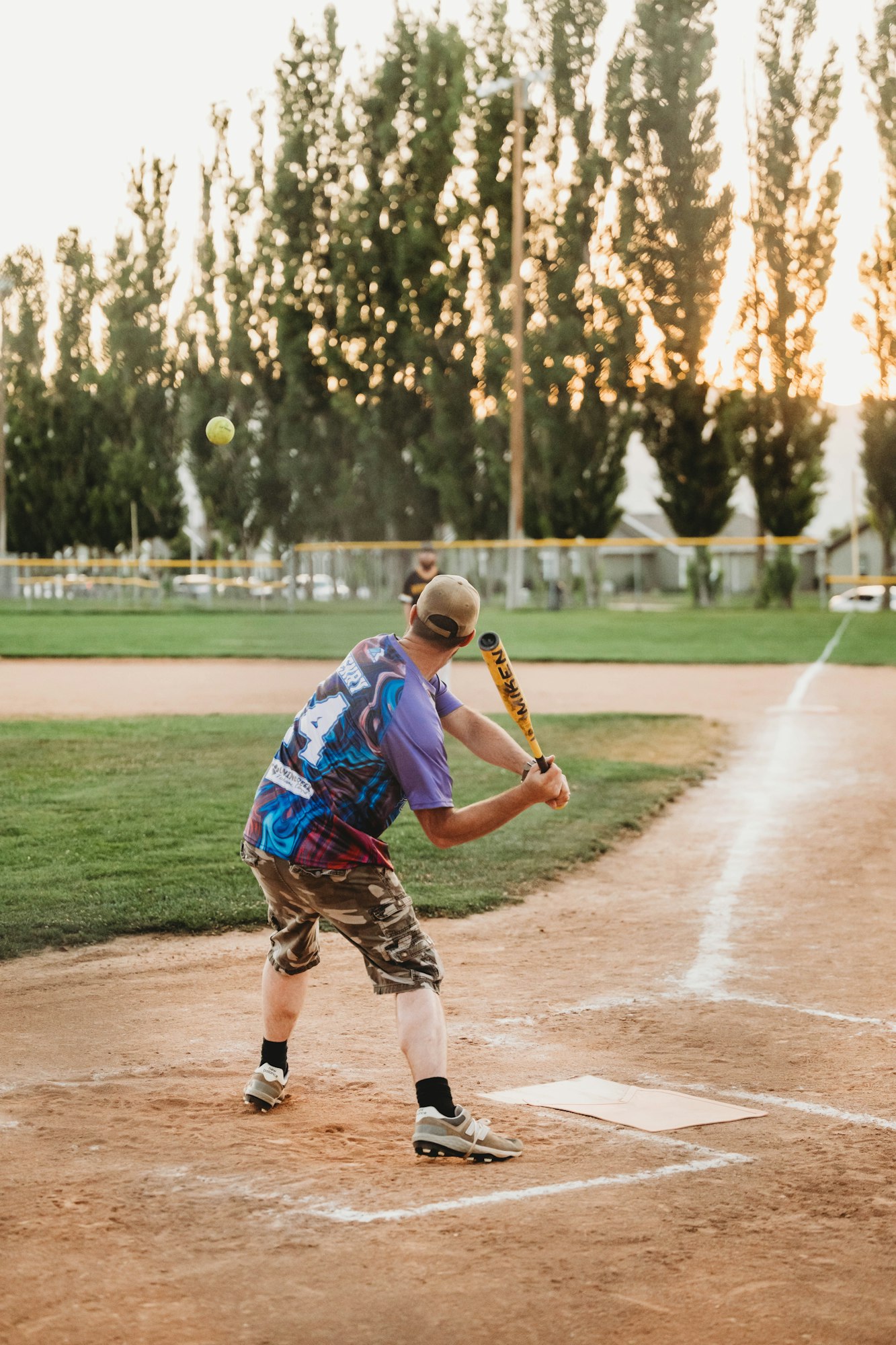 A person playing softball, swinging at a pitch, with trees and a fence in the background.