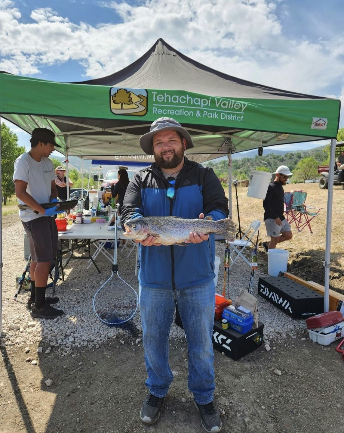 A man holding a large fish with a fishing net and outdoor gear in the background under a canopy.