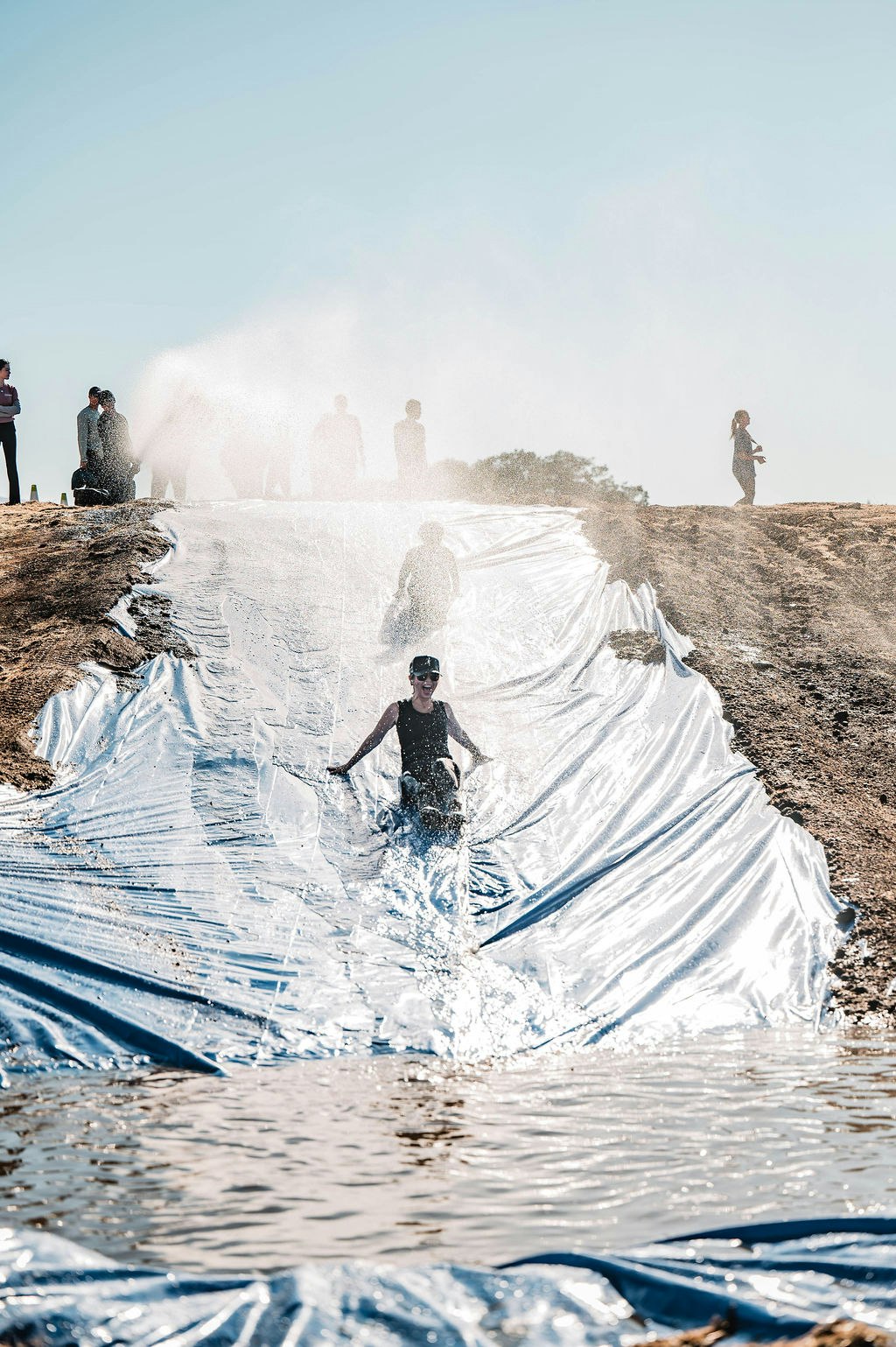 Person sliding down a water slide made of tarp on a hill, splashing into water, with others standing in the background.