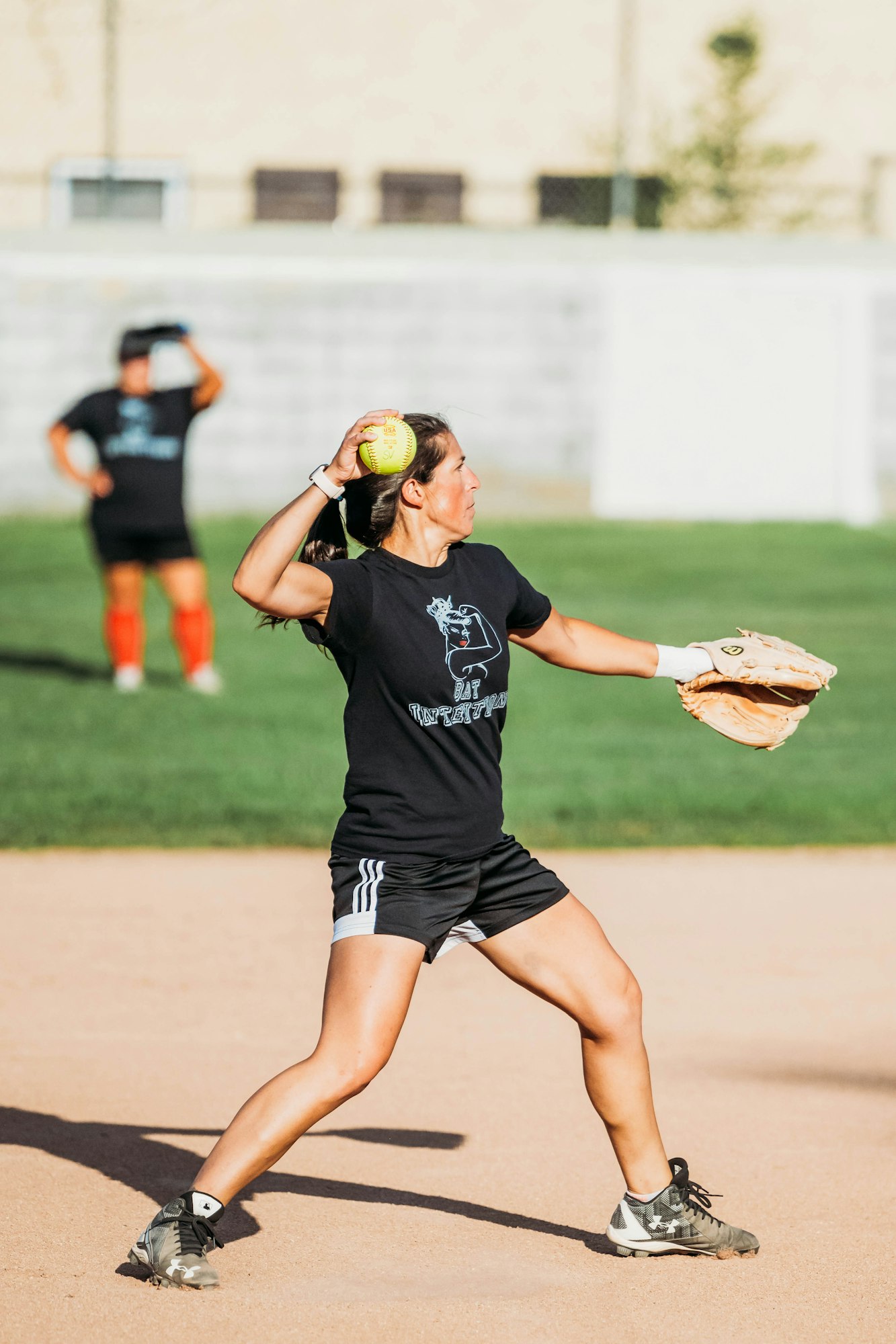 A player throwing a softball on a field, another player in the background.