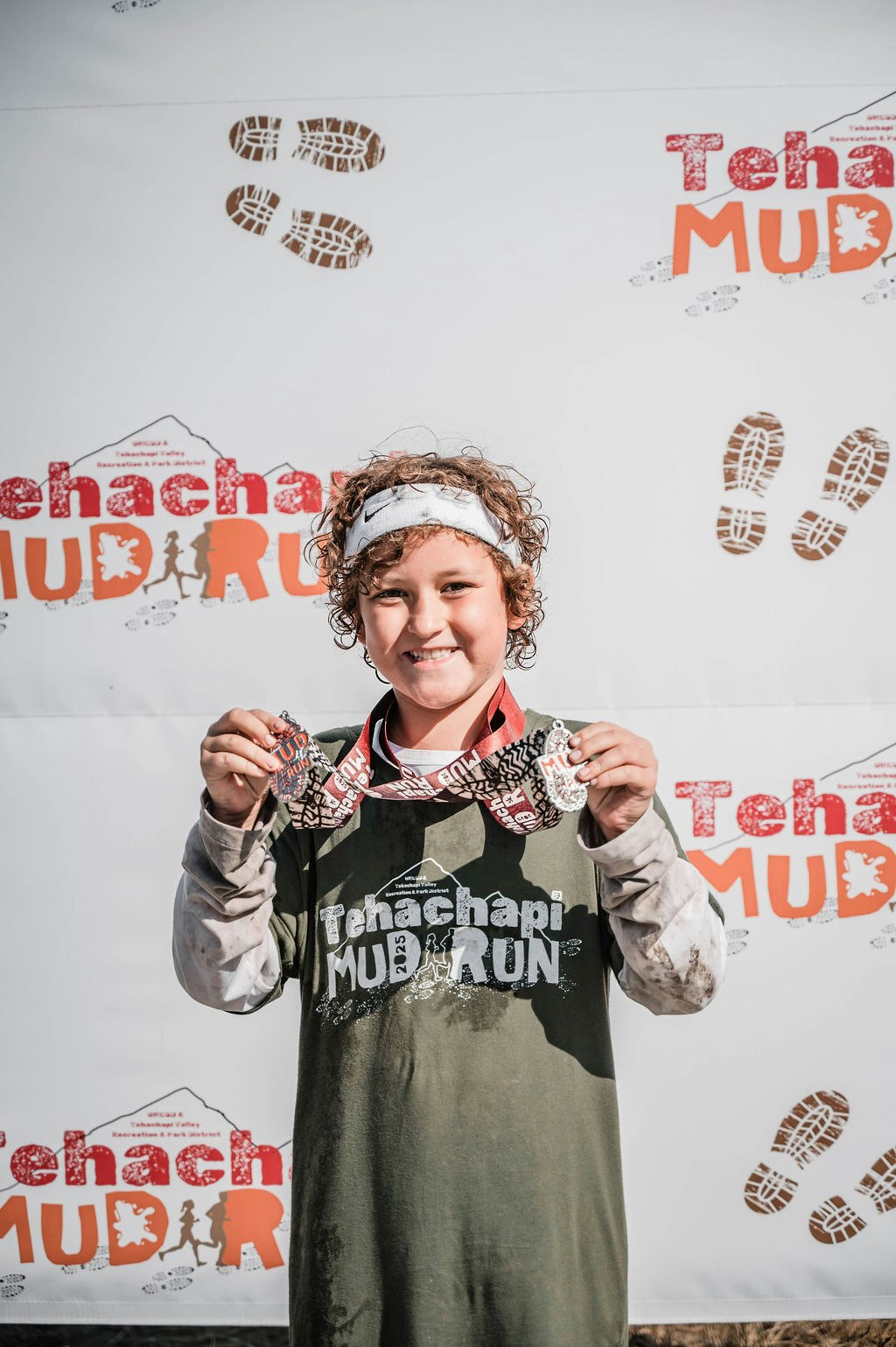A person smiling, holding medals, wearing a "Tehachapi Mud Run" shirt and headband, with a mud run backdrop behind them.