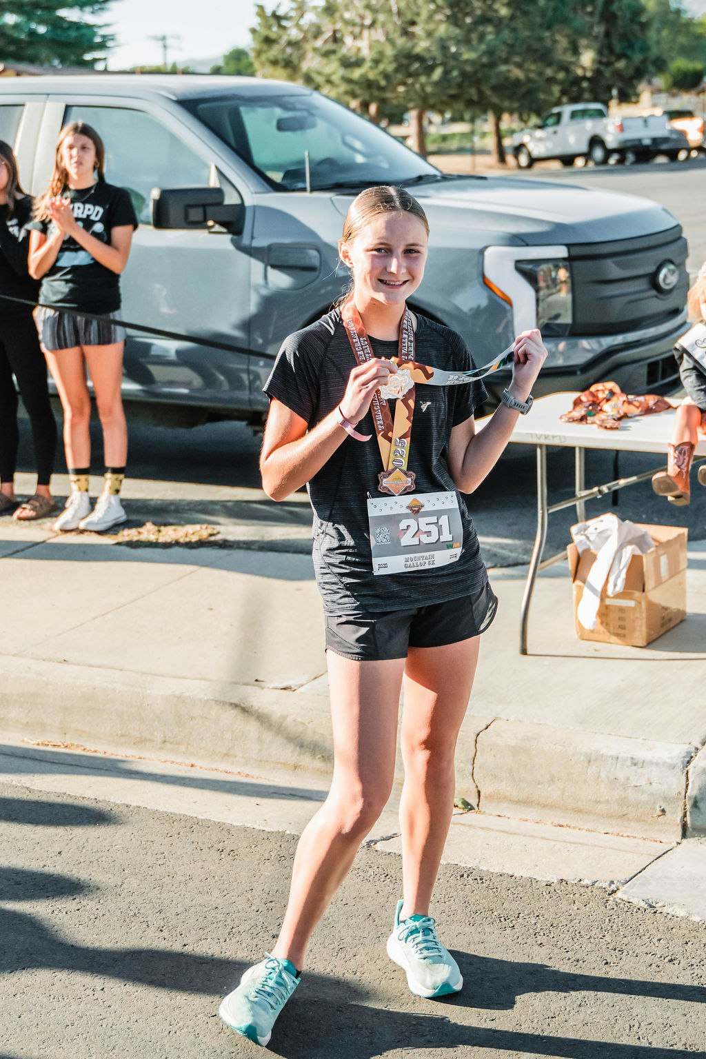 A young person holding a medal and wearing a race bib, standing outdoors with others applauding in the background.