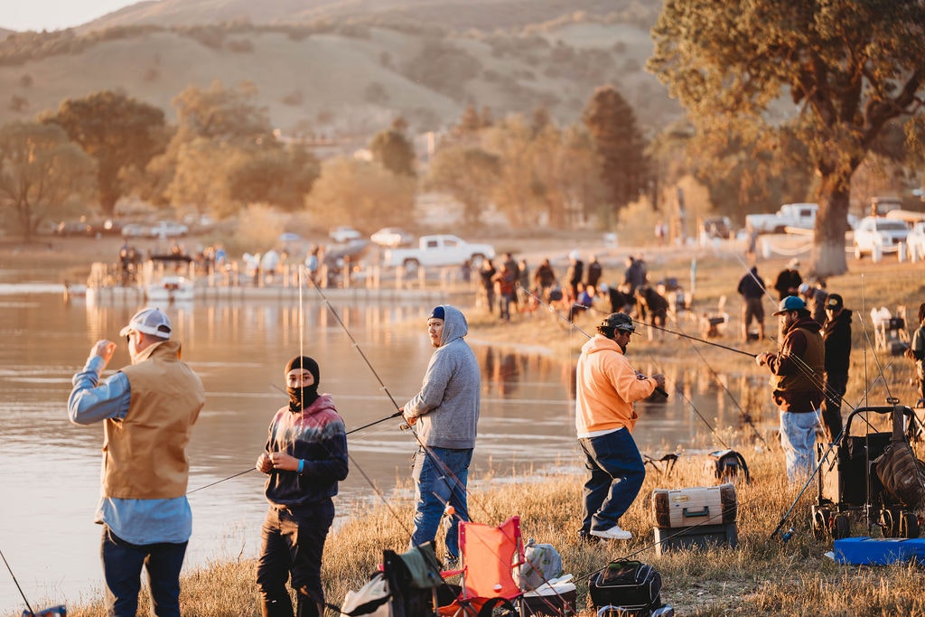 People fishing by a lake at sunset, with camping tents and cars in the background.