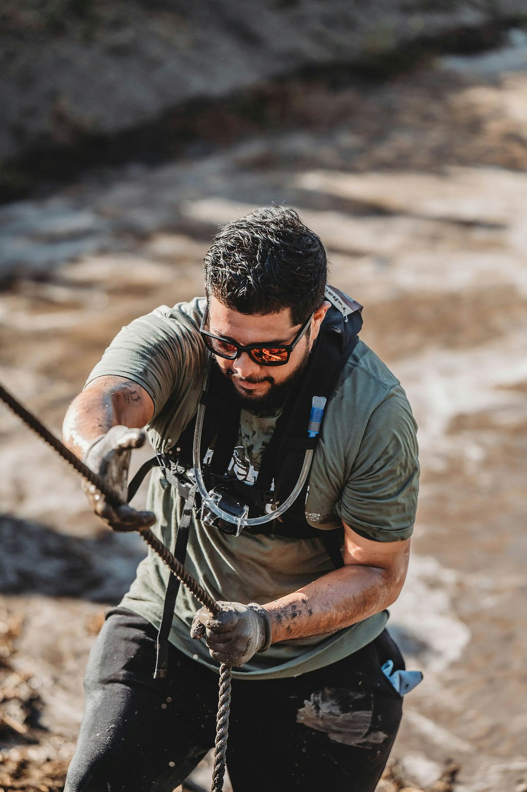 A man climbing a muddy slope with a rope, wearing sunglasses and a hydration pack.