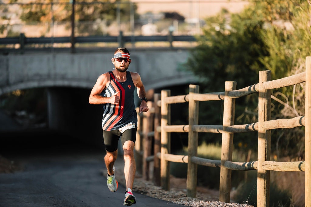 A man in sports attire is running on a pathway beside a wooden fence with a bridge in the background.