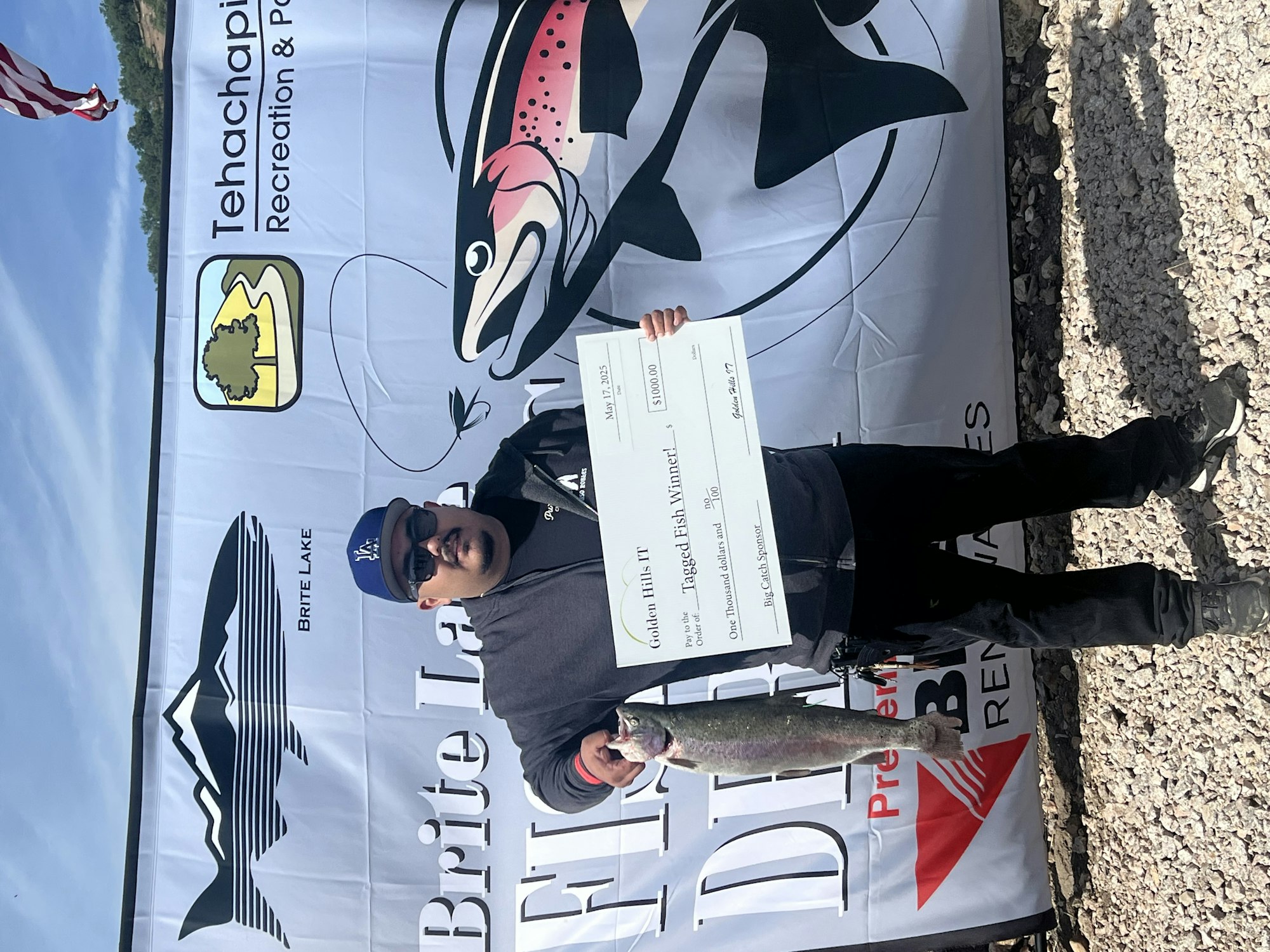 A person holding a fish and a large check, with a fishing event banner in the background.