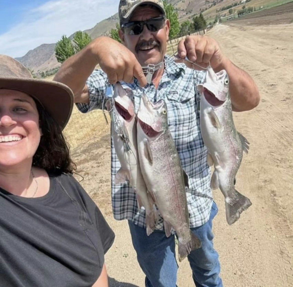 Two people smiling with a string of fish; one person holding the fish, the other taking a selfie, outdoors.