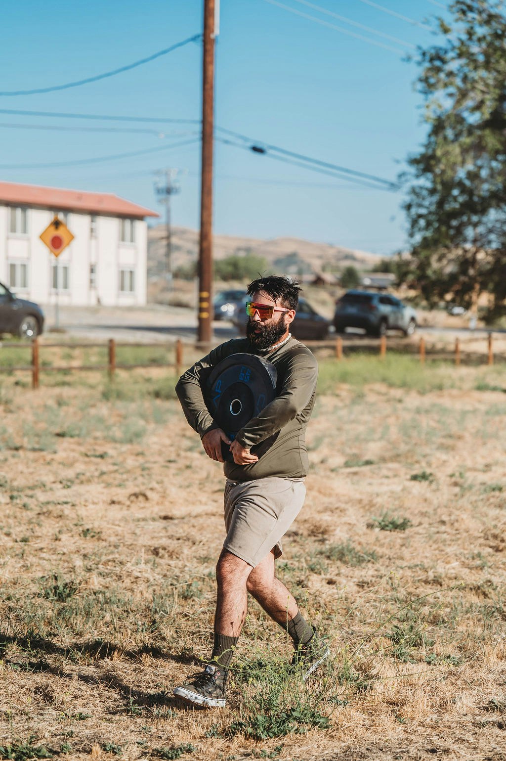 A bearded man in sunglasses carries a weight plate across a grassy field.