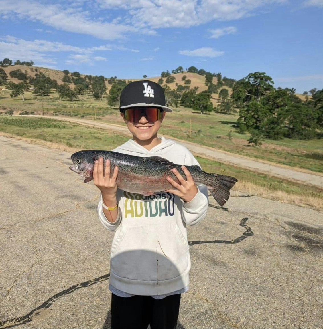 A child wearing sunglasses and a cap proudly holding a large fish with a scenic hilly landscape in the background.