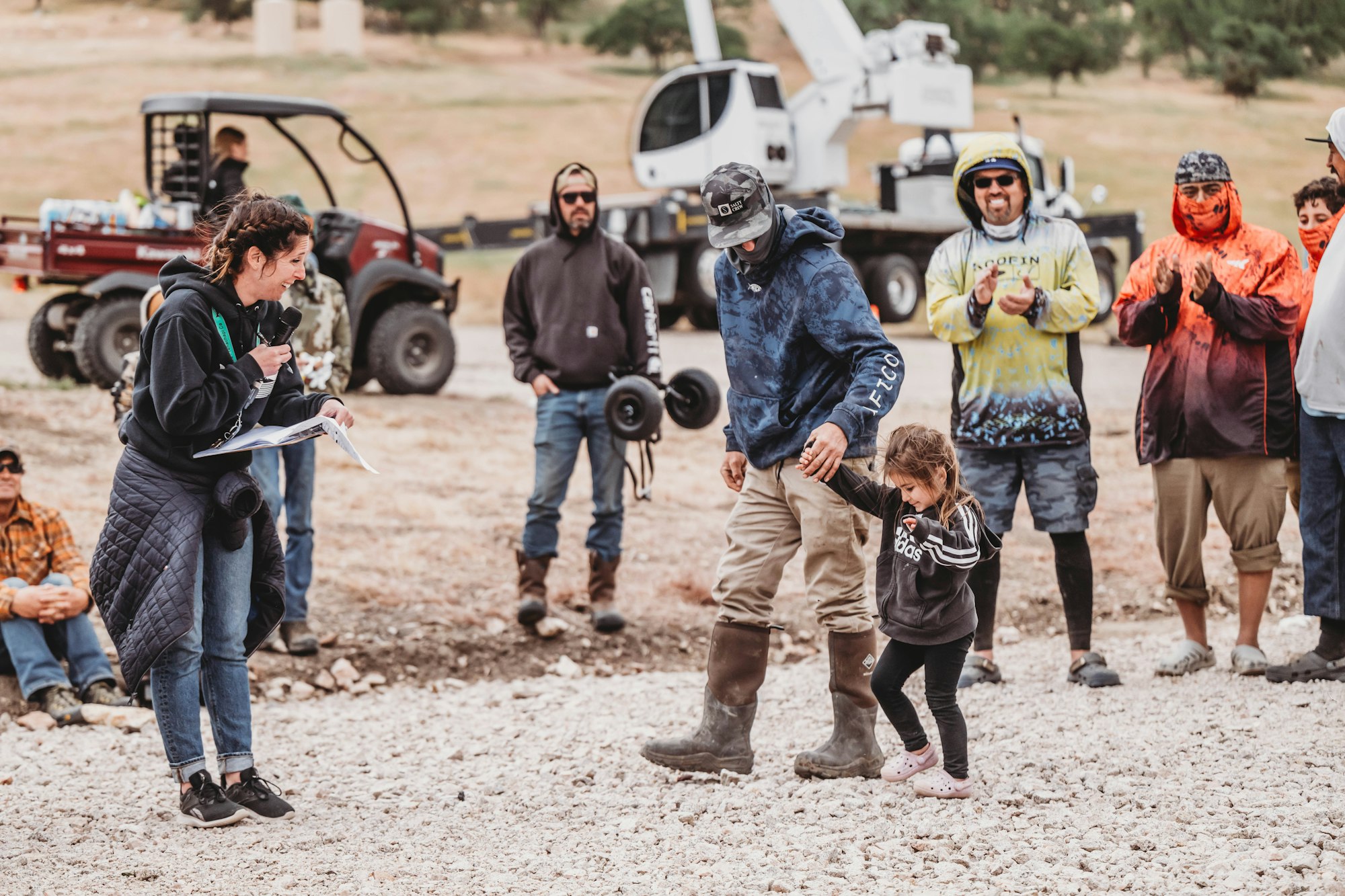 A group of people outdoors, including a man and child walking hand in hand. Others are watching, some clapping, next to vehicles.