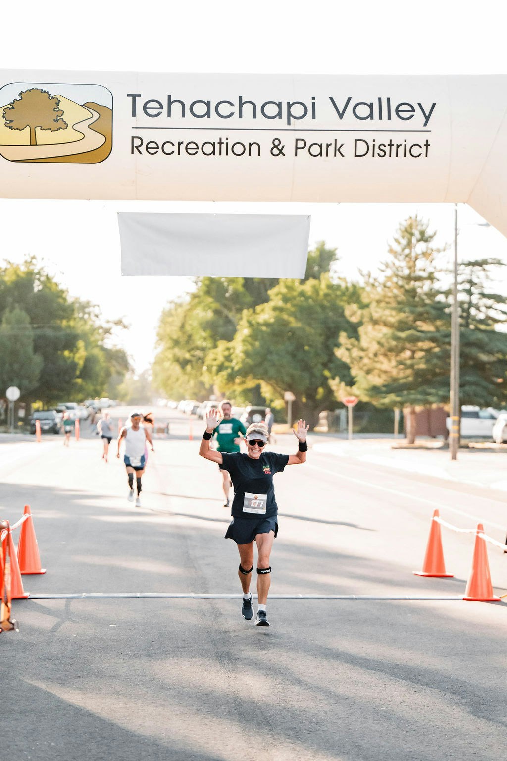 A runner finishes a race under a "Tehachapi Valley Recreation & Park District" banner, raising arms in celebration.